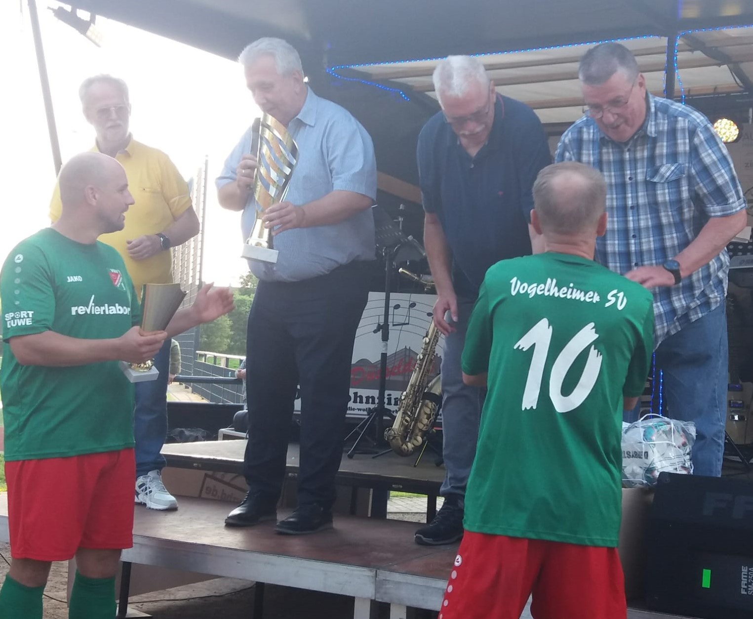 [{"clang_id":1,"value":"Eine Gruppe von Männern auf einer Bühne. Zwei in grünen Fußballtrikots mit Nummern, drei weitere in Hemden und Hosen, einige spielen Blasinstrumente."}]