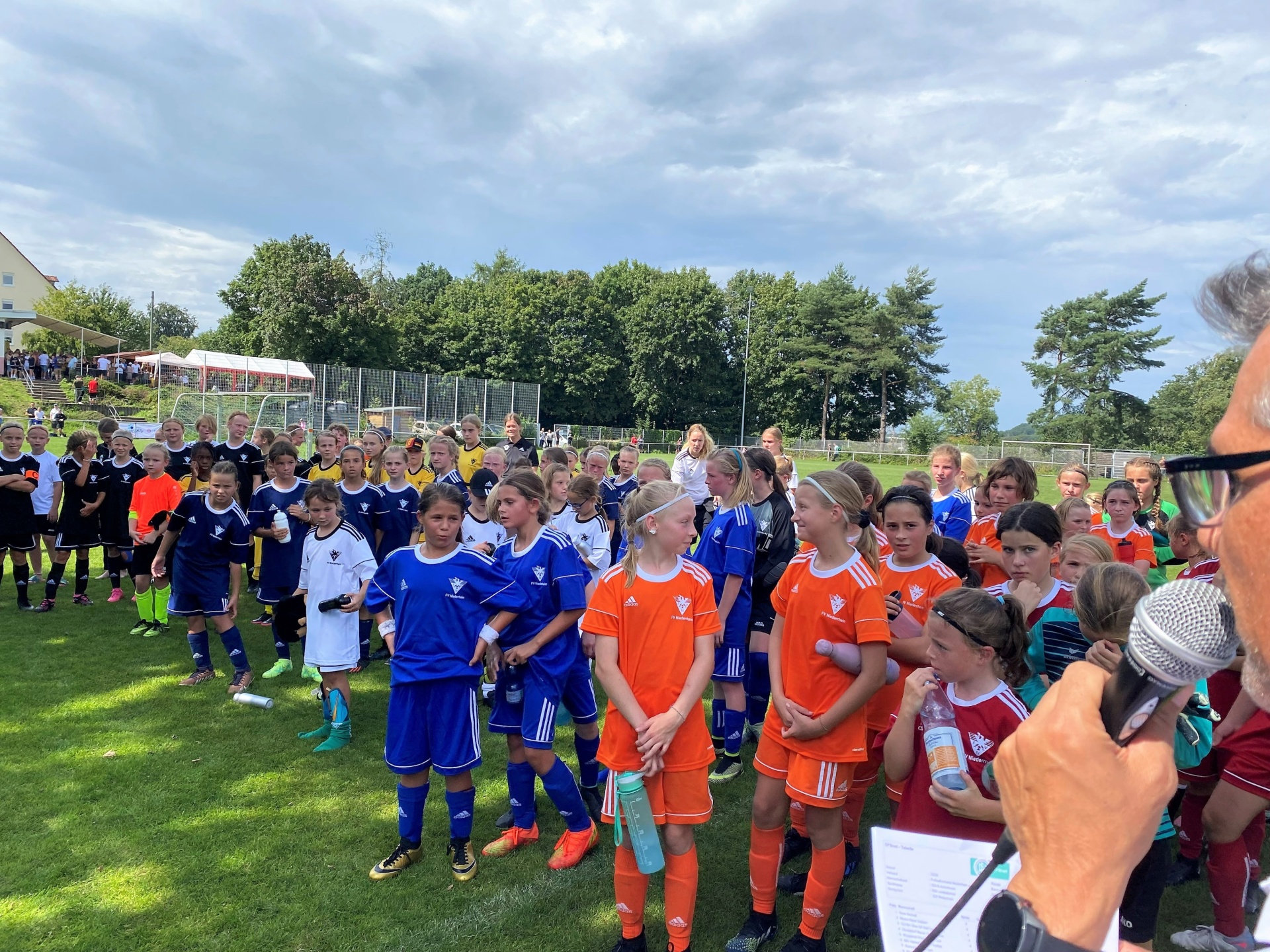 [{"clang_id":1,"value":"Fußballspielerinnen in blau und orange stehen auf einem Rasenplatz und hören einer Person mit Mikrofon zu. Im Hintergrund sind Zuschauer und Bäume zu sehen."}]