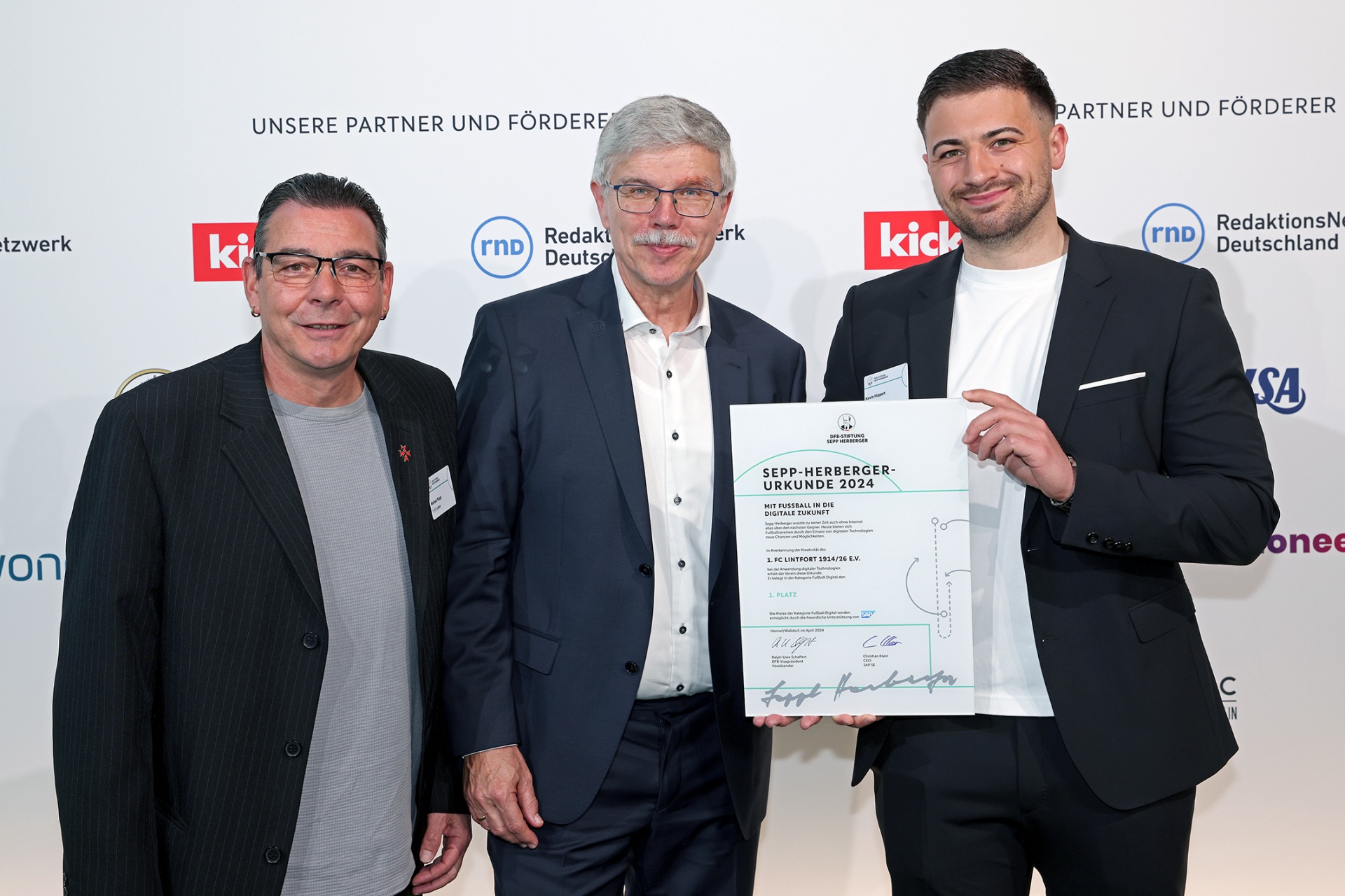 [{"clang_id":1,"value":"Drei Männer in Anzügen stehen lächelnd vor einem Banner. Der Mann rechts hält eine Urkunde mit einem Fußballmotiv. Im Hintergrund Logos verschiedener Organisationen."}]