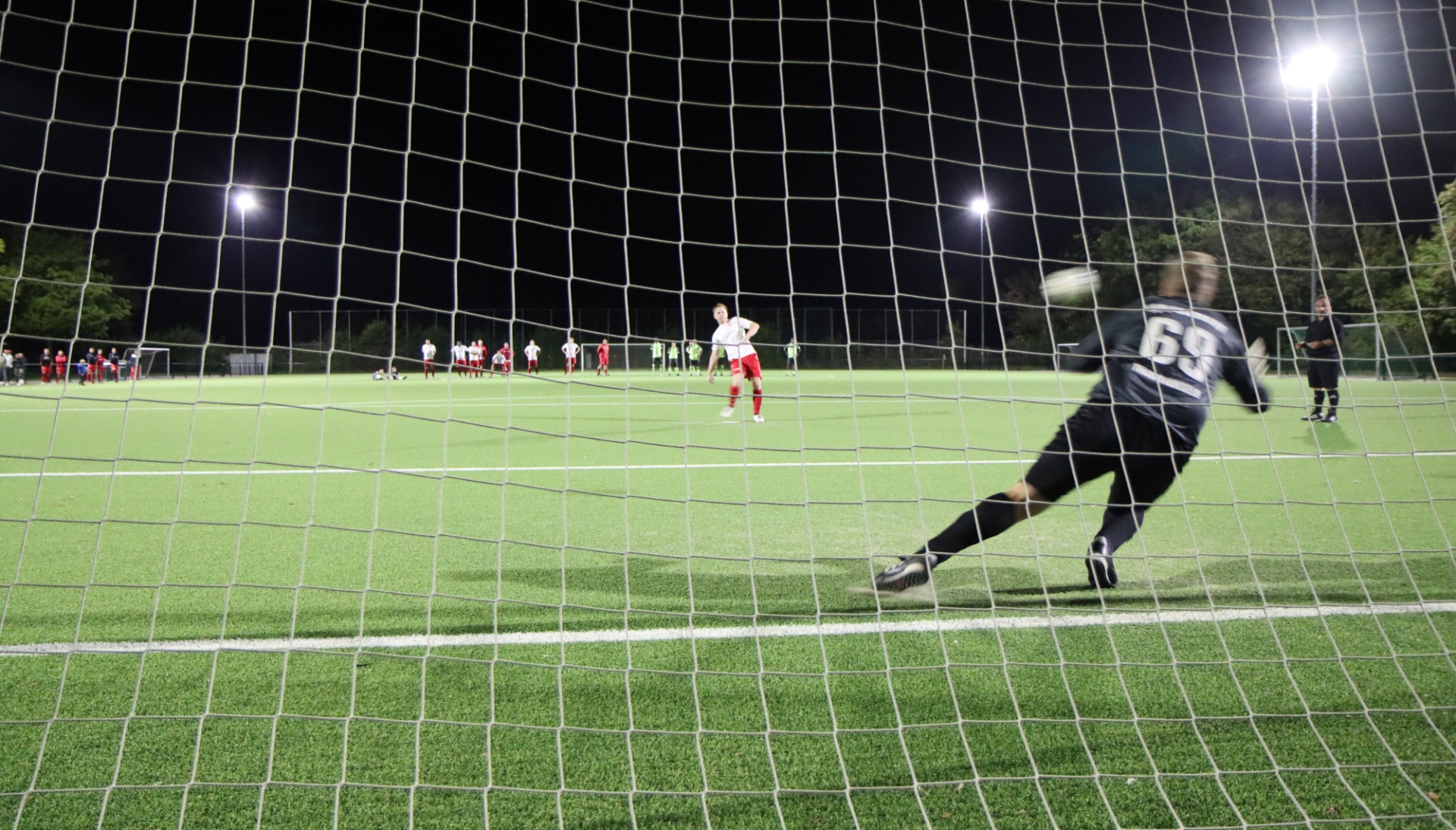 [{"clang_id":1,"value":"Fußballtorwart springt zur Seite, um einen Schuss abzuwehren. Im Hintergrund sind weitere Spieler auf einem grünen Rasenfeld bei Nacht zu sehen."}]