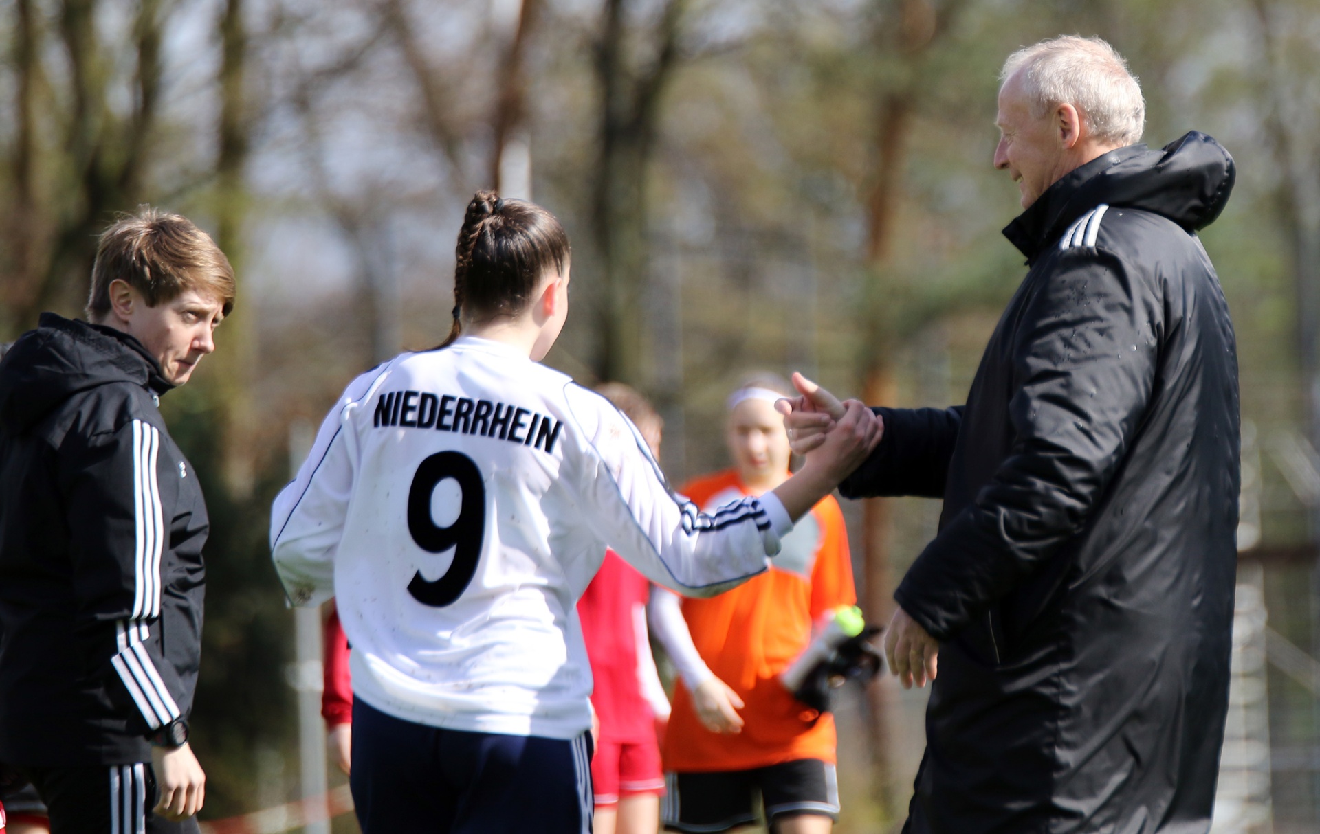 [{"clang_id":1,"value":"Fußballfeld. Zwei Trainer sprechen mit einem Jugendspieler (Trikotnummer 9, Schriftzug \"NIEDERRHEIN\"). Im Hintergrund weitere Spieler in Sportkleidung."}]