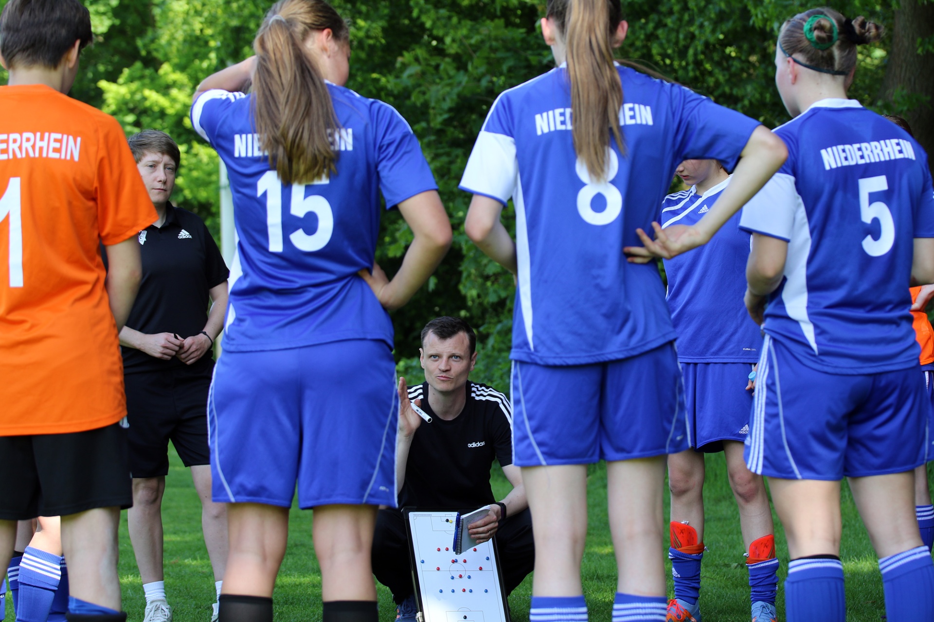 [{"clang_id":1,"value":"Fußballteam in blauen Trikots und orangefarbenem Torwart-Shirt steht im Kreis um Trainer mit Taktiktafel. Hintergrund: Grünfläche."}]