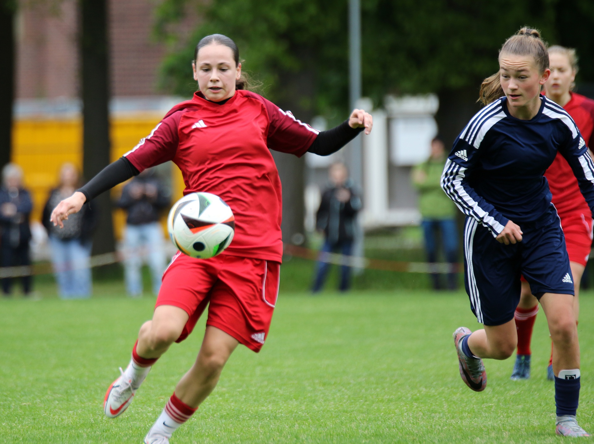 [{"clang_id":1,"value":"Zwei Fußballspielerinnen auf Grünfeld. Eine trägt rot, dribbelt den Ball, die andere in Dunkelblau läuft ihr entgegen. Im Hintergrund Zuschauer und Gebäude."}]