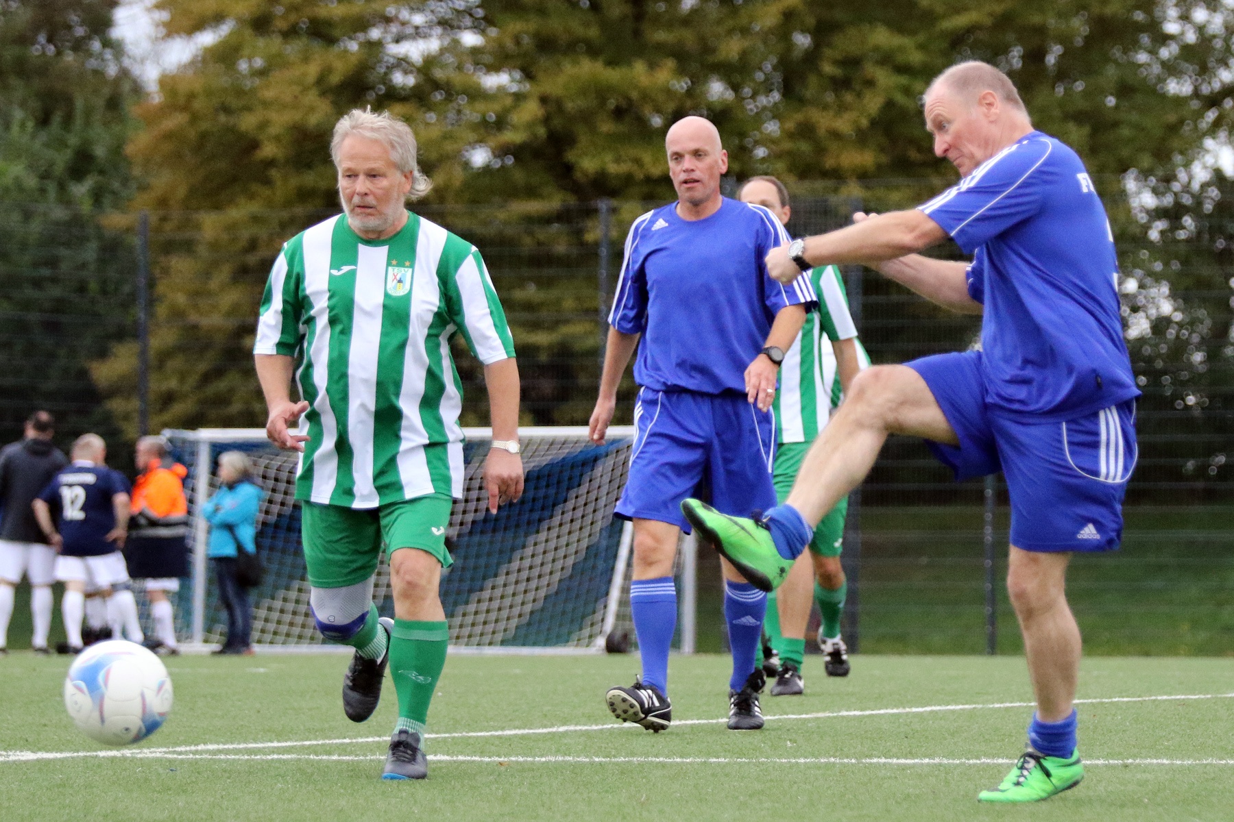 [{"clang_id":1,"value":"Fußballspiel mit älteren Herren auf Rasenplatz. Zwei Spieler im Vordergrund, einer in grün-weißem, der andere in blauem Trikot, verfolgen den Ball. Zuschauer im Hintergrund."}]