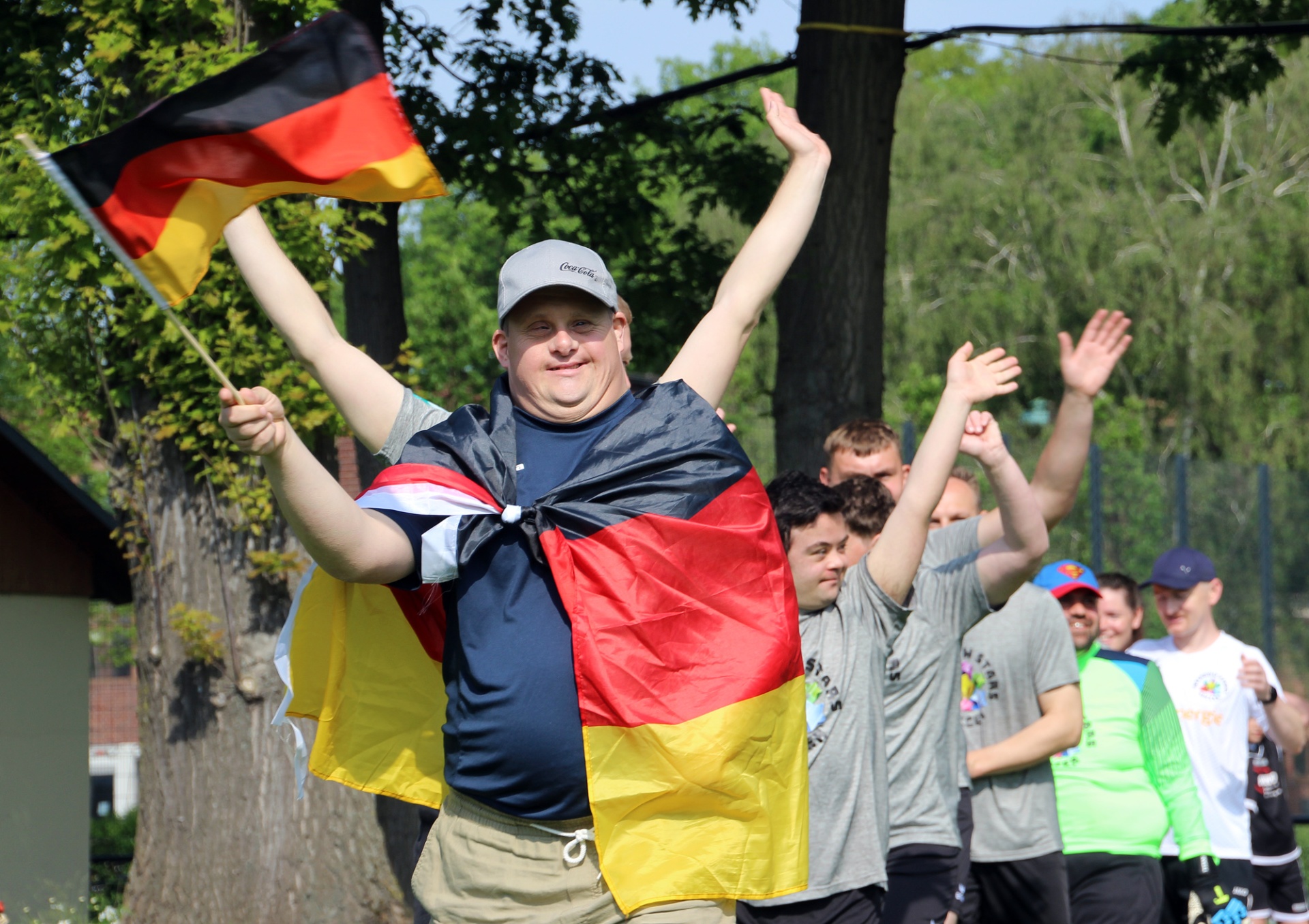 [{"clang_id":1,"value":"Mann mit Deutschlandflagge und Flaggentuch, Arme hoch. Im Hintergrund Gruppe Menschen, Bäume und Gebäude."}]