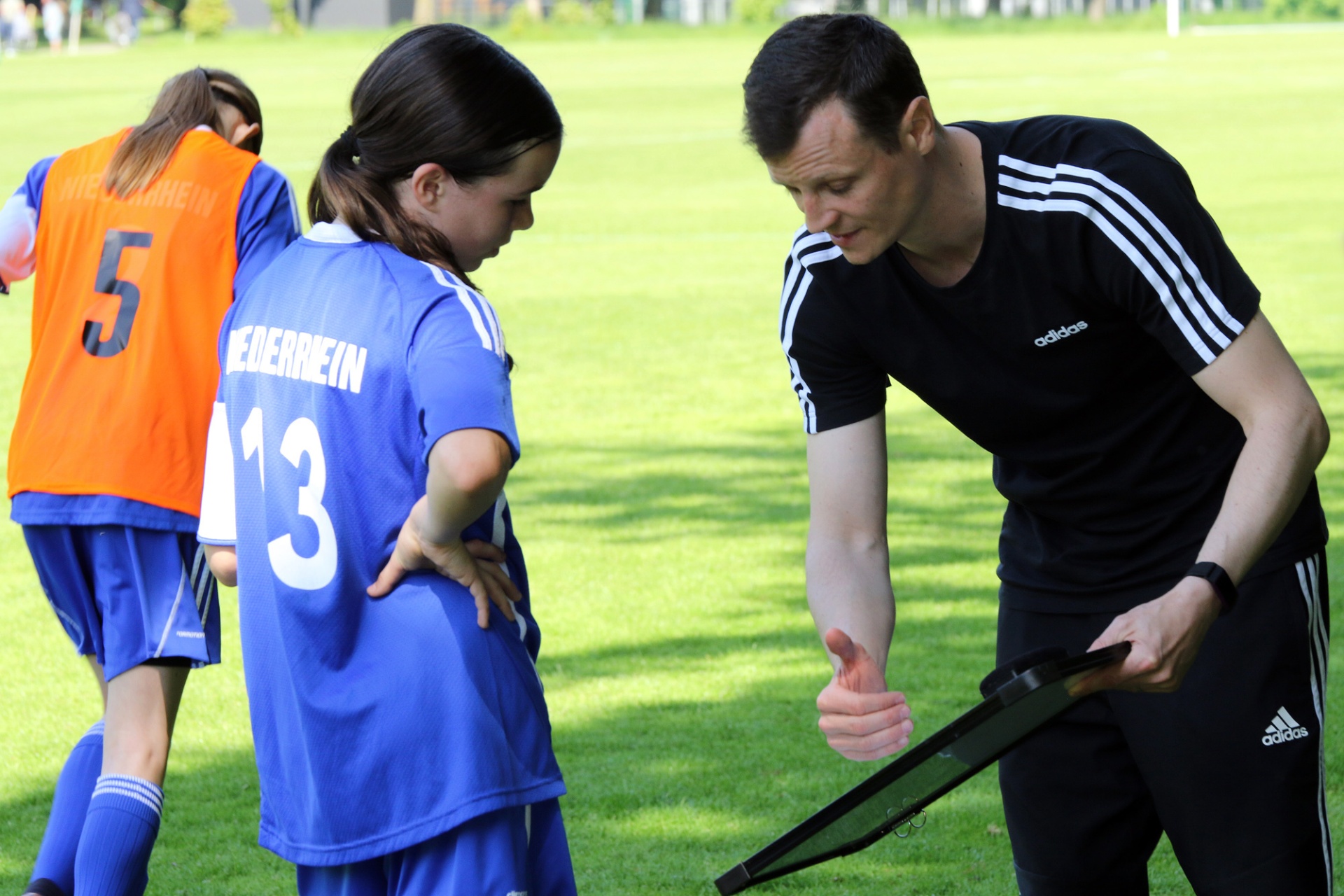 [{"clang_id":1,"value":"Fußballtrainerin/Trainer erklärt junge Spielerin (Trikotnummer 13) etwas auf dem Grün. Im Hintergrund weitere Spielerinnen in orangefarbenen Trikots."}]