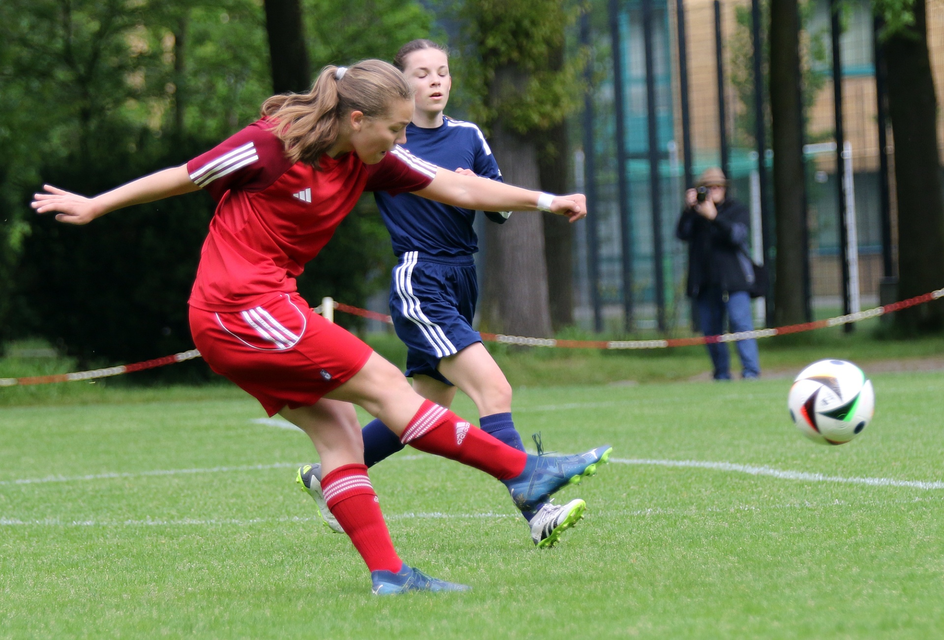 [{"clang_id":1,"value":"Zwei Fußballspielerinnen auf Grün. Eine in Rot schießt, die andere in Blau verteidigt. Im Hintergrund Gebäude und Zuschauer."}]