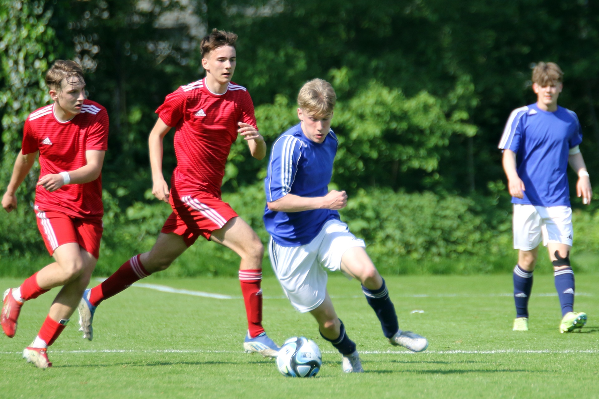 [{"clang_id":1,"value":"Fußballspiel: Zwei Spieler in Rot verfolgen einen Spieler in Blau mit Ball auf grünem Rasen. Im Hintergrund ein weiterer Spieler in Blau."}]