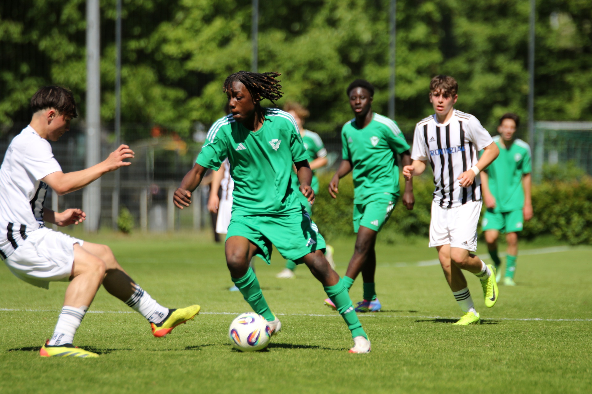 [{"clang_id":1,"value":"Fußballspiel: Jugendlicher mit Ball dribbelt, verfolgt von Gegenspielern in Schwarz-Weiß und Teamkollegen in Grün. Rasenplatz an sonnigem Tag."}]