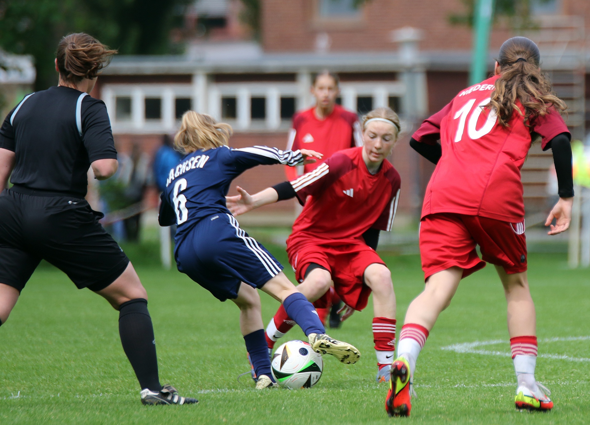 [{"clang_id":1,"value":"Fußballspiel: Drei Spielerinnen kämpfen um den Ball auf grünem Rasen. Eine in rot, eine in blau, eine in schwarz. Im Hintergrund Gebäude und weitere Spieler."}]