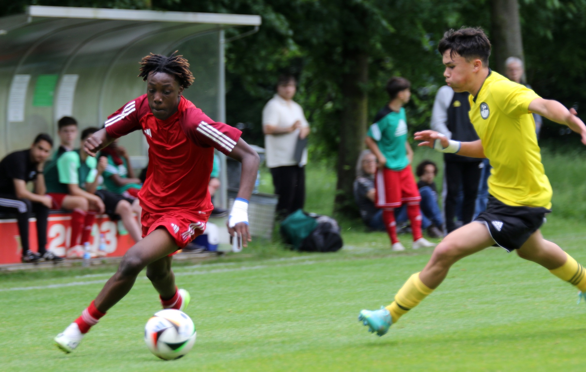 [{"clang_id":1,"value":"Zwei Fußballspieler im Spiel: Links in Rot dribbelt den Ball, rechts in Gelb versucht, ihn zu erobern. Hintergrund: Zuschauer und Bäume."}]