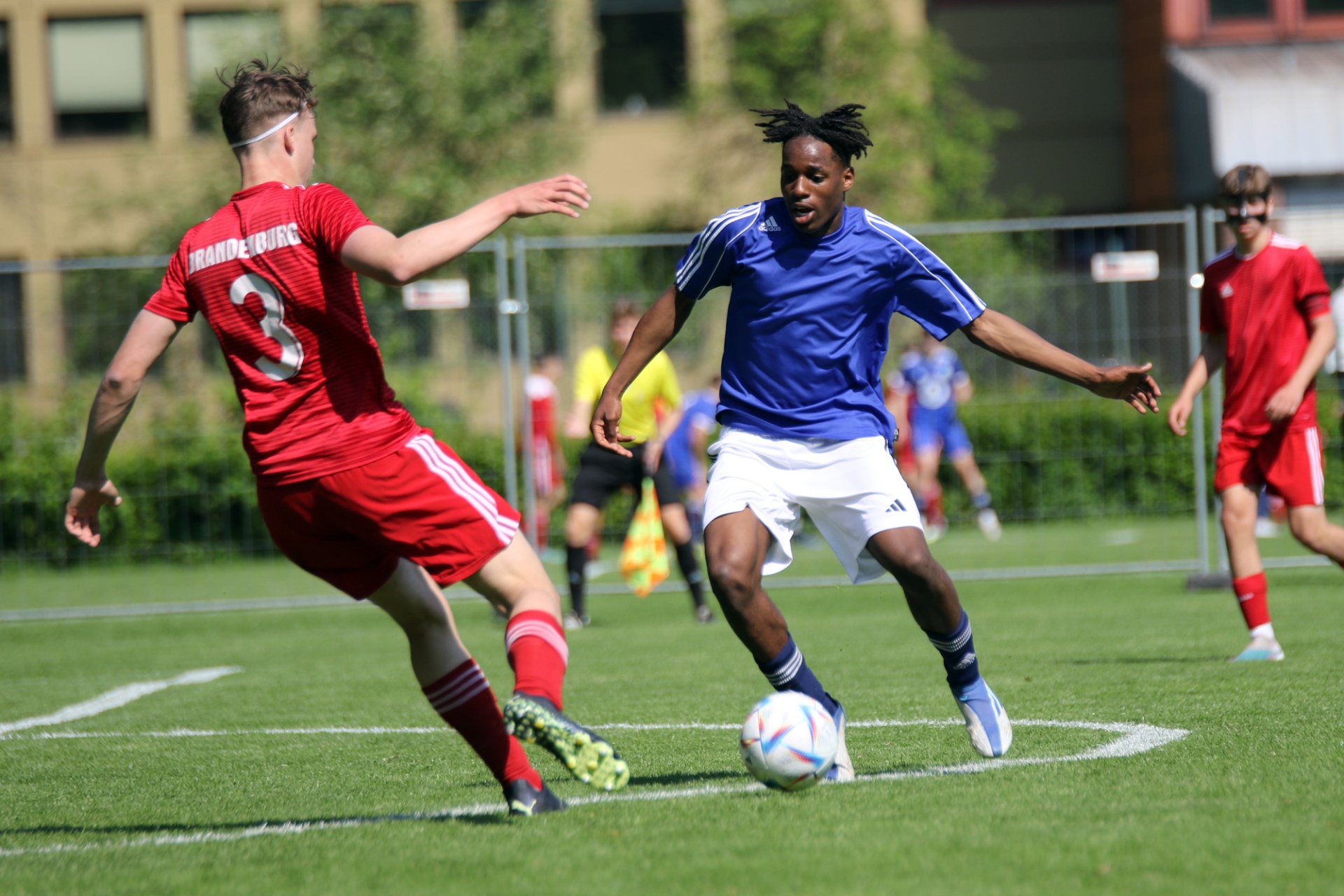 [{"clang_id":1,"value":"Zwei Fußballspieler im Spiel: Links in Rot, rechts in Blau dribbelt den Ball. Im Hintergrund weitere Spieler und ein Gebäude."}]