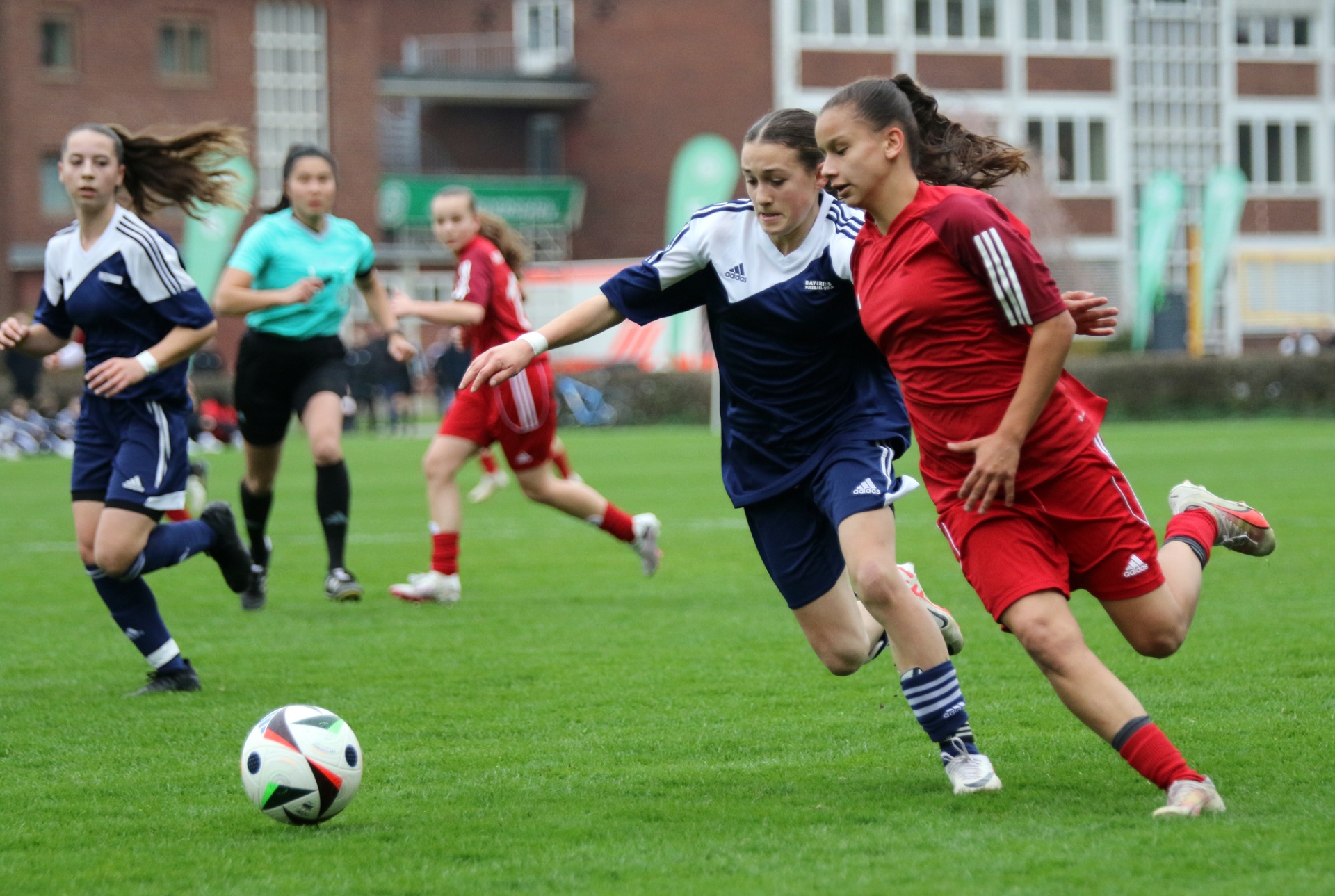 [{"clang_id":1,"value":"Fußballspiel: Zwei junge Frauen dribbeln den Ball, weitere Spielerinnen im Hintergrund auf dem grünen Rasen. Gebäude im Hintergrund."}]