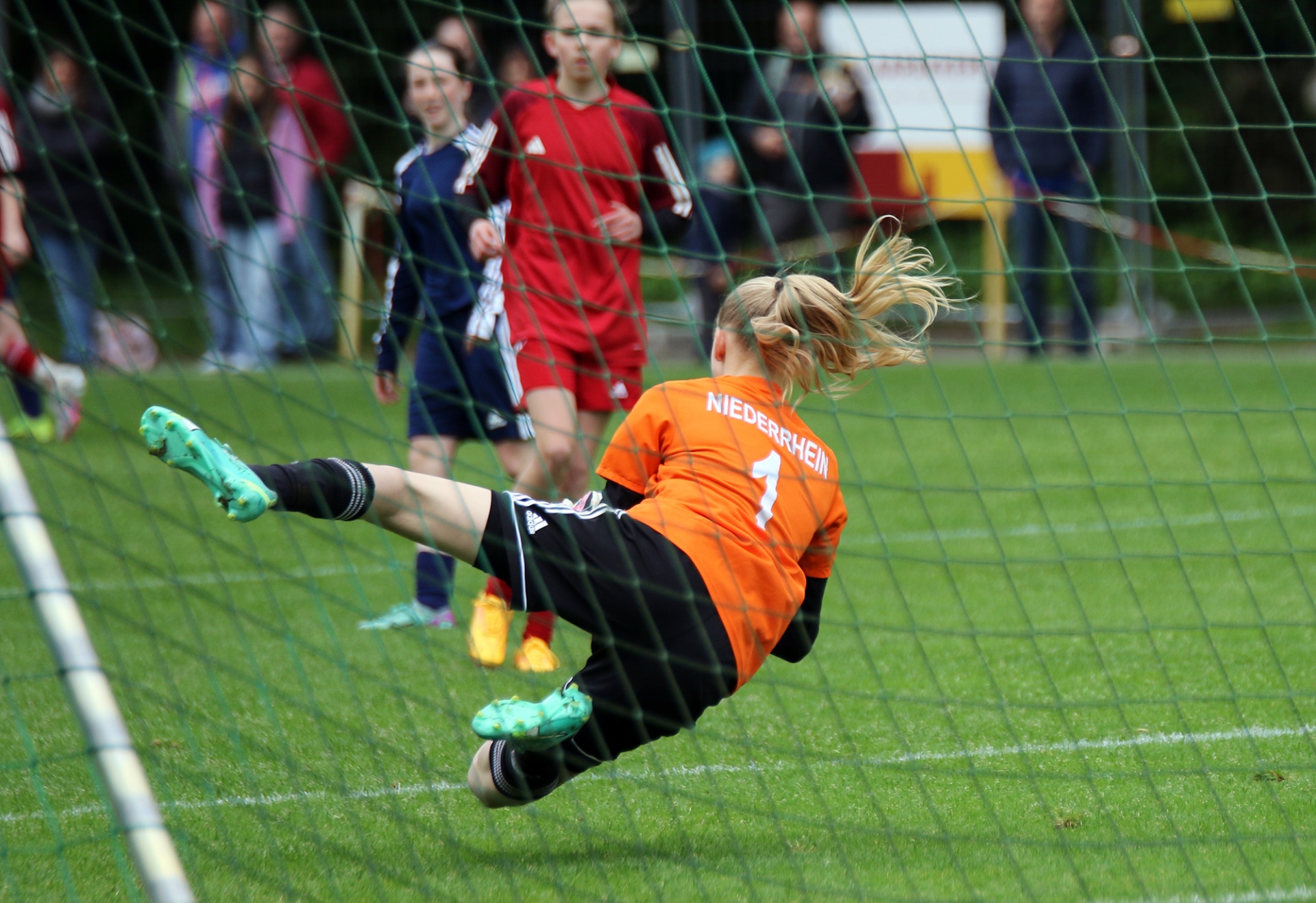 [{"clang_id":1,"value":"Fußballtorwartin in orangefarbenem Trikot springt im Gras nach einem Ball. Im Hintergrund stehen Spieler und Zuschauer."}]