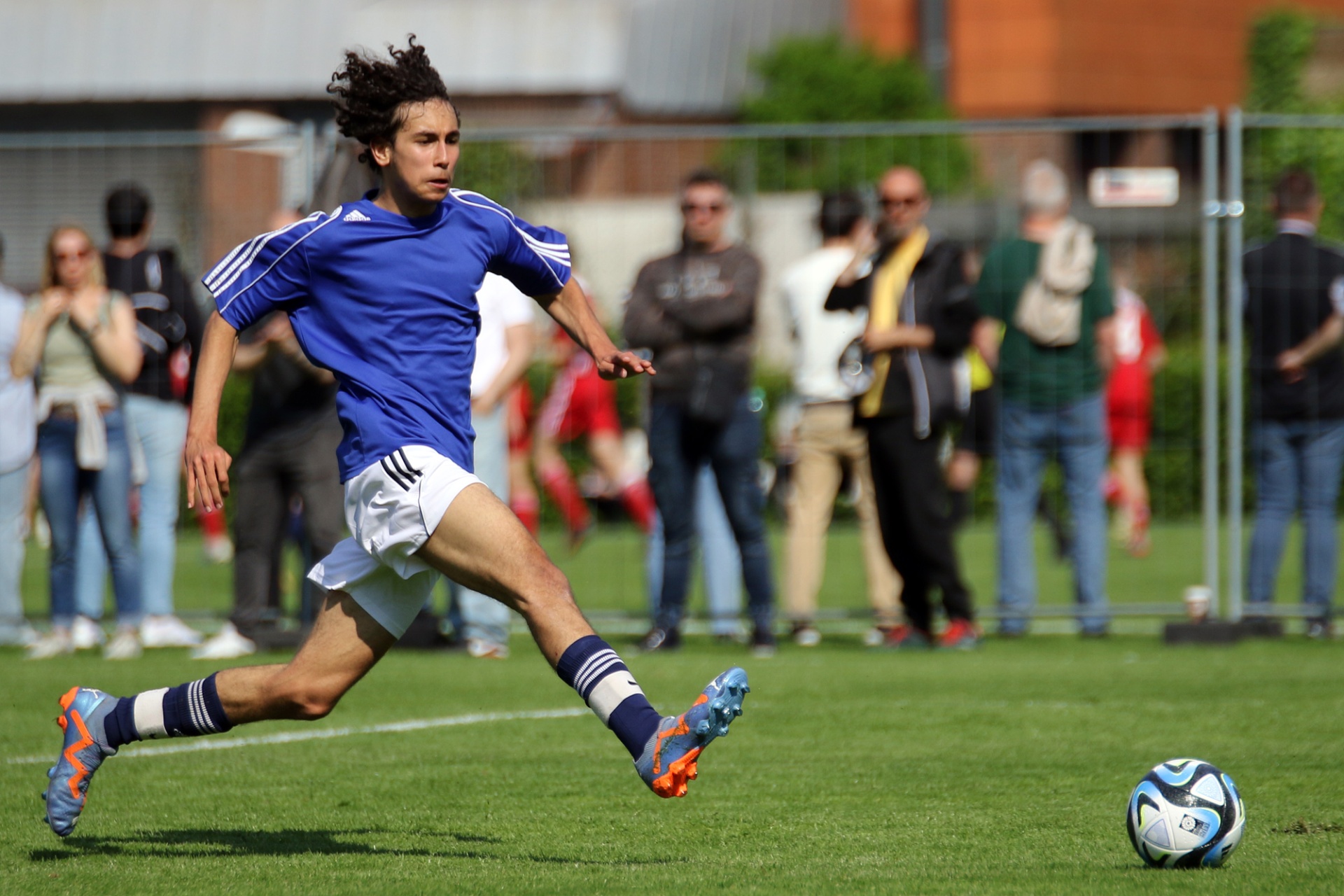 [{"clang_id":1,"value":"Fußballspieler mit blauer Uniform und weißer Hose schießt auf ein Fußballfeld. Im Hintergrund Zuschauer und Gebäude."}]
