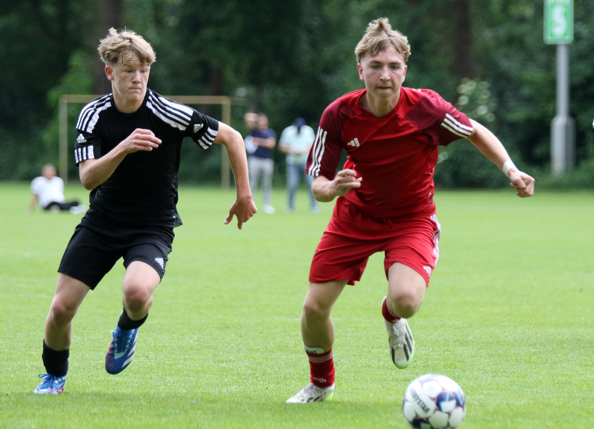 [{"clang_id":1,"value":"Zwei junge Fußballspieler rennen auf einem grünen Rasenfeld. Links trägt der Spieler schwarze Kleidung, rechts rote. Im Hintergrund weitere Personen und ein Fußballtor."}]