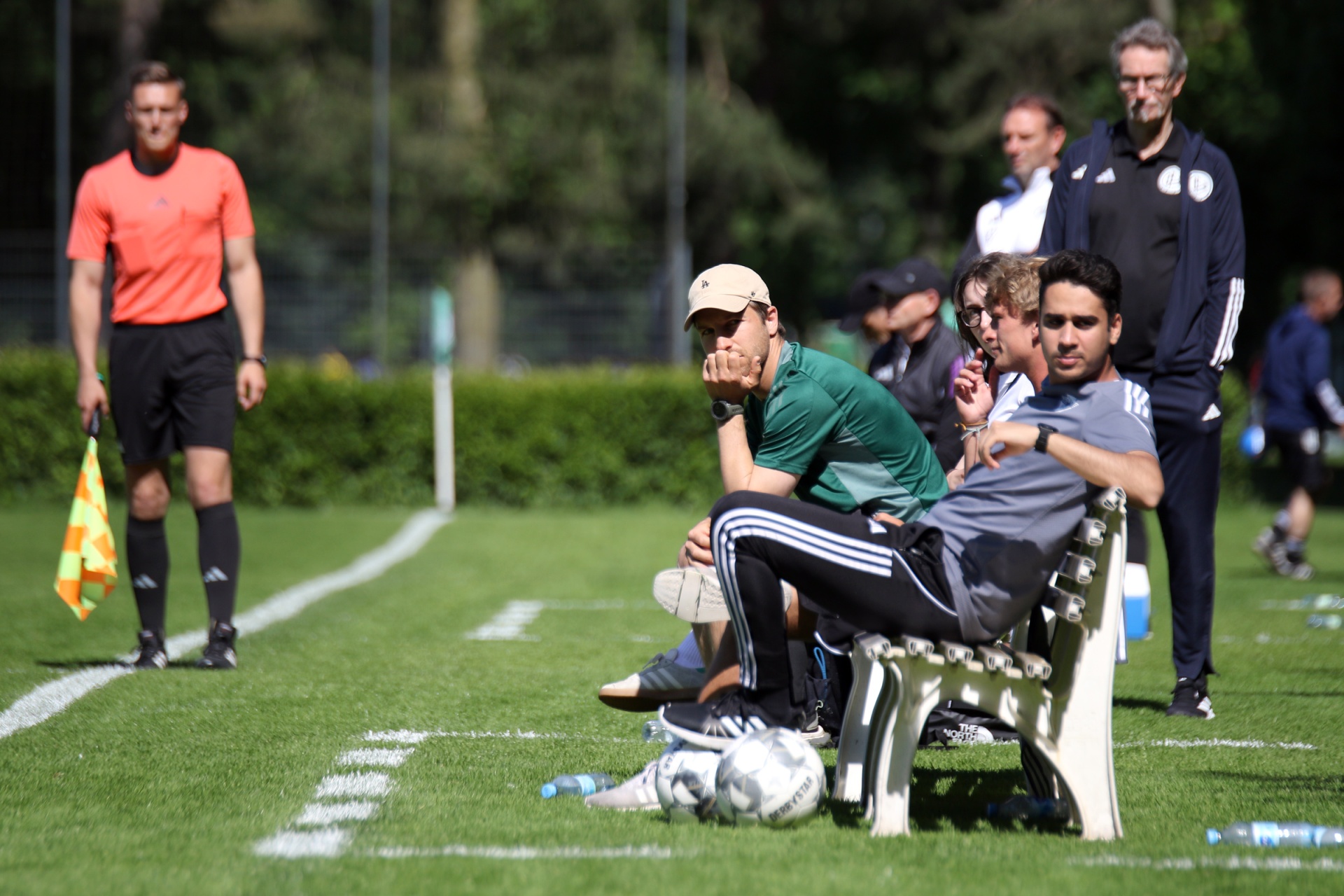 [{"clang_id":1,"value":"Fußballfeld mit Schiedsrichter, mehreren Männern auf einer Bank und Fußbällen davor. Bäume im Hintergrund."}]