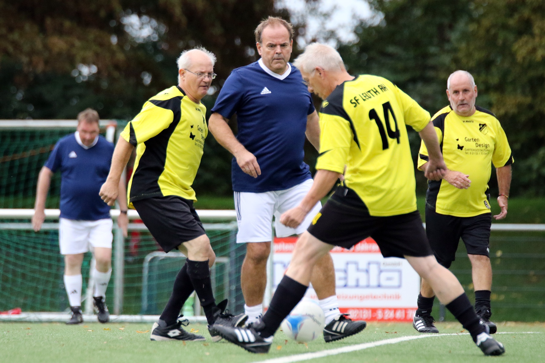 [{"clang_id":1,"value":"Fußballspiel mit vier älteren Männern auf einem Rasenplatz. Zwei tragen gelbe Trikots, einer blau, einer steht mit verschränkten Armen im Hintergrund."}]