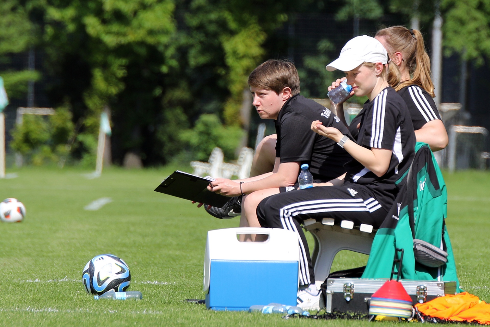 [{"clang_id":1,"value":"Zwei Personen sitzen auf einer Bank am Fußballplatz. Der Mann schreibt auf ein Clipboard, die Frau trinkt Wasser. Fußbälle und Trainingsmaterial liegen im Gras."}]