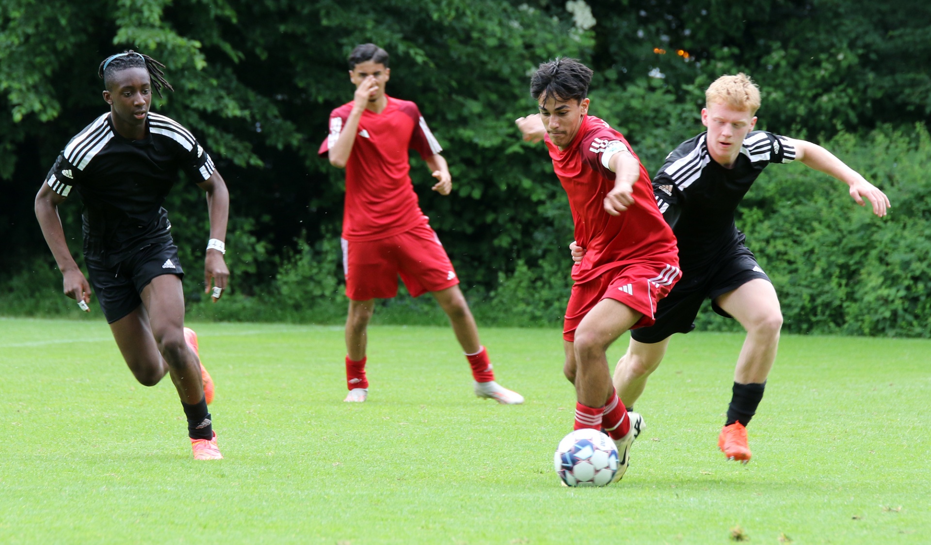 [{"clang_id":1,"value":"Vier Fussballspieler auf grünem Rasen. Zwei in roten, zwei in schwarzen Trikots. Ein Spieler dribbelt den Ball. Bäume im Hintergrund."}]