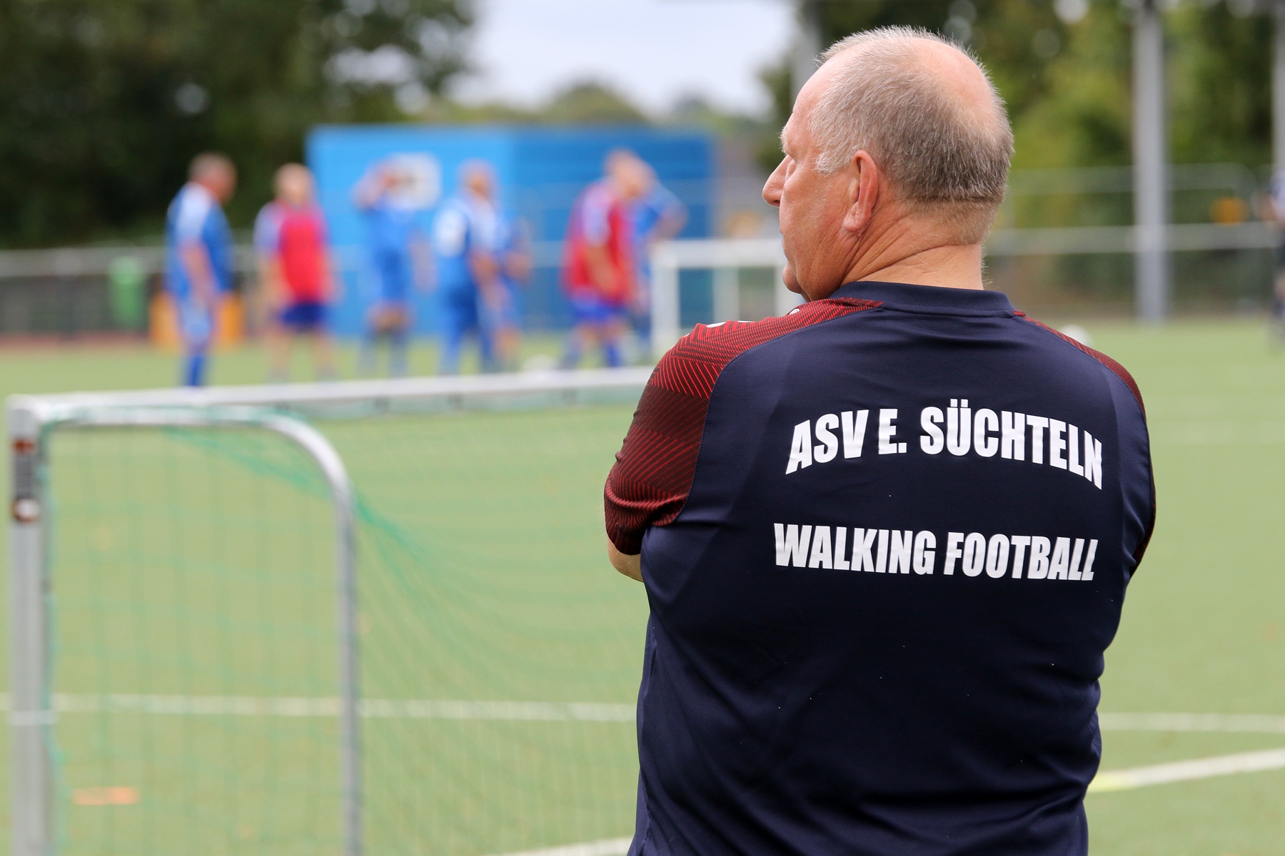 [{"clang_id":1,"value":"Mann mit blauem Trikot \"ASV E. Süchtel Walking Football\" steht auf einem Fußballfeld und beobachtet ein Spiel mit rot- und blau gekleideten Spielern."}]