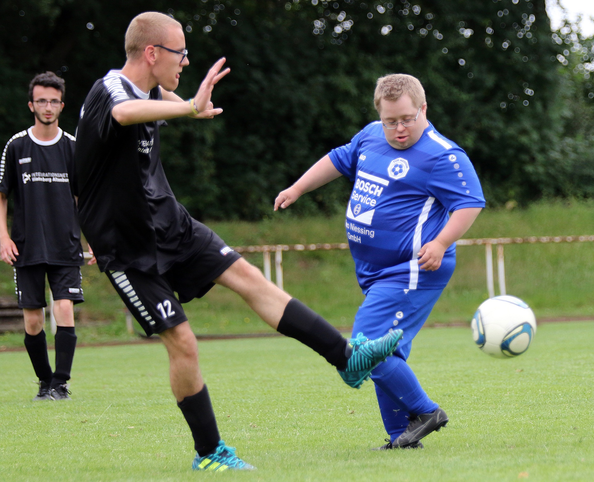 [{"clang_id":1,"value":"Zwei Fußballspieler auf grünem Rasen. Links ein Mann mit Trikot Nr. 12, rechts ein Mann mit Down-Syndrom in blauem Trikot, beide beim Spielzug. Im Hintergrund weitere Personen und Büsche."}]