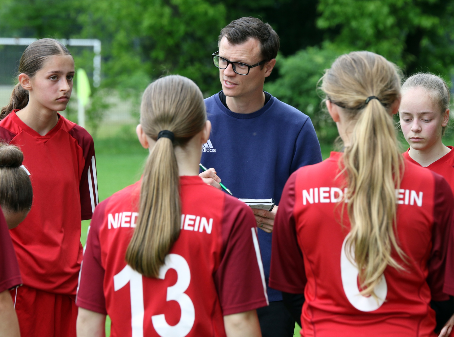 [{"clang_id":1,"value":"Fußballtrainer bespricht Taktik mit vier jugendlichen Spielerinnen auf dem Rasen. Alle tragen rote Trikots mit weißen Nummern und dem Schriftzug \"NIEDER\"."}]