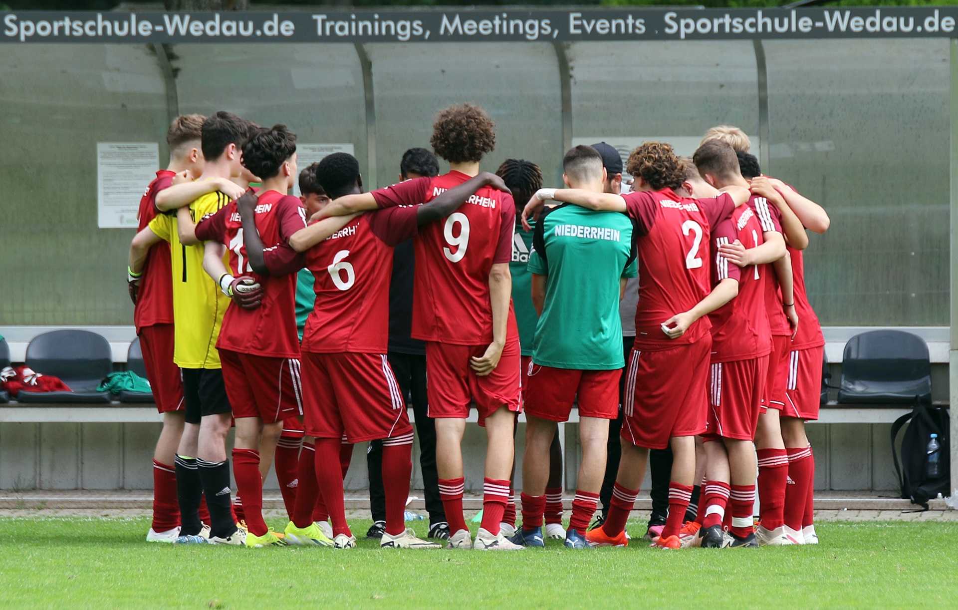 [{"clang_id":1,"value":"Fußballmannschaft in roten Trikots im Kreis vereint auf grünem Rasen. Einige Spieler tragen Nummern (2, 6, 9). Im Hintergrund eine Bank und Werbebanner."}]