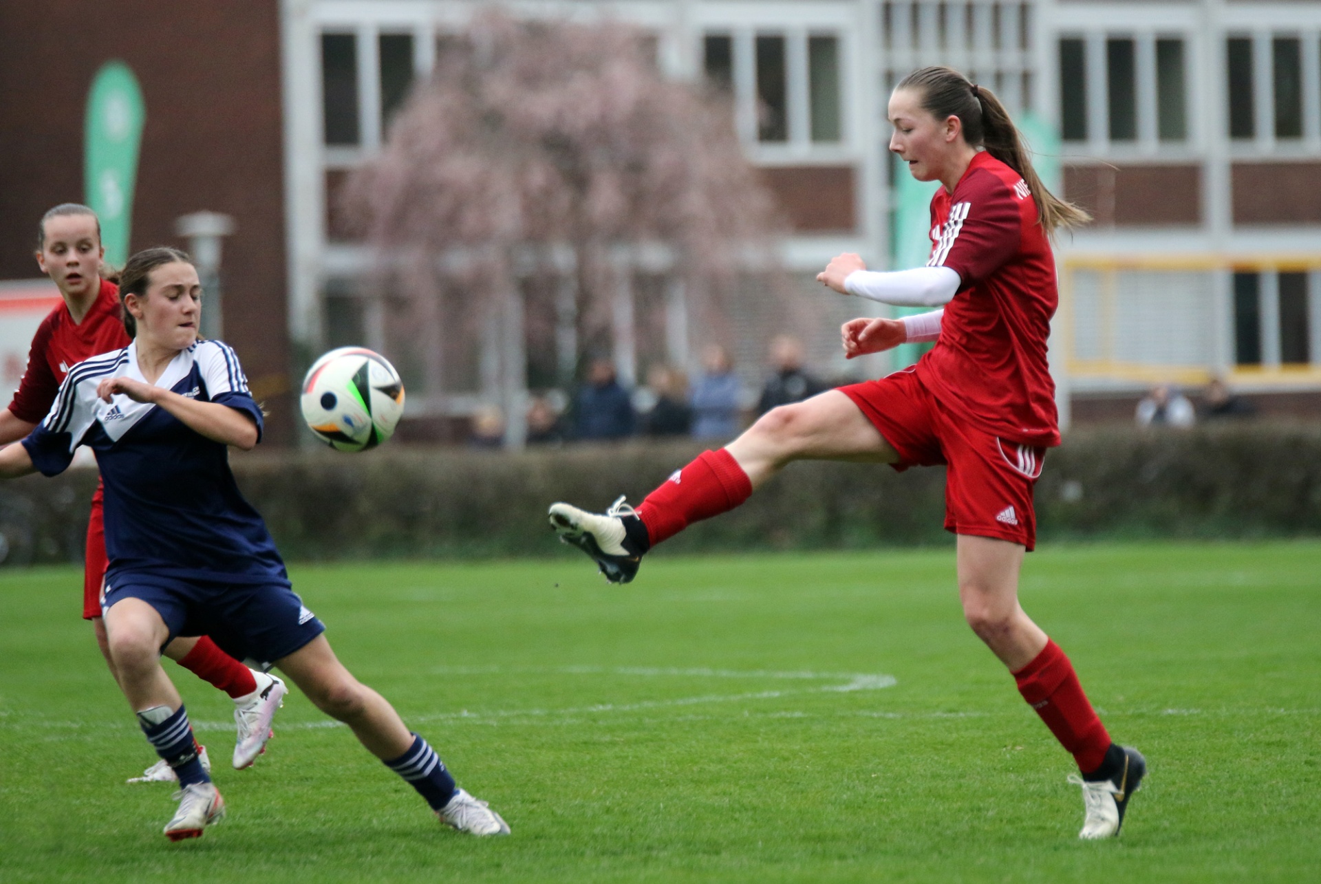 [{"clang_id":1,"value":"Zwei Fußballspielerinnen auf grünem Rasen. Eine in Rot schießt auf den Ball, die andere in Blau versucht ihn zu blocken. Im Hintergrund Gebäude und Bäume."}]