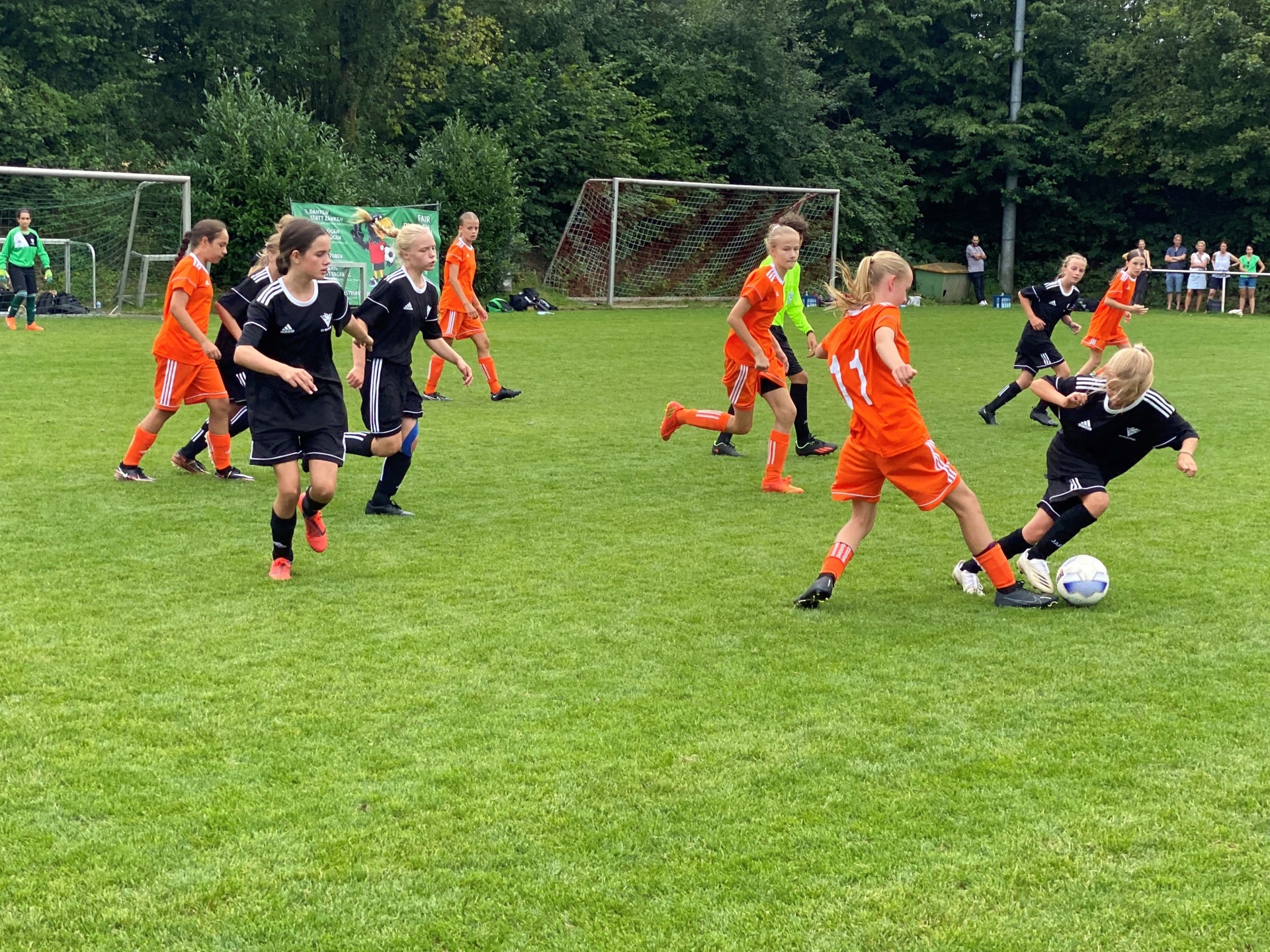 [{"clang_id":1,"value":"Fußballspiel: Spieler in schwarzen und orangefarbenen Trikots kämpfen um den Ball auf einem grünen Rasenplatz. Im Hintergrund Bäume und Zuschauer."}]