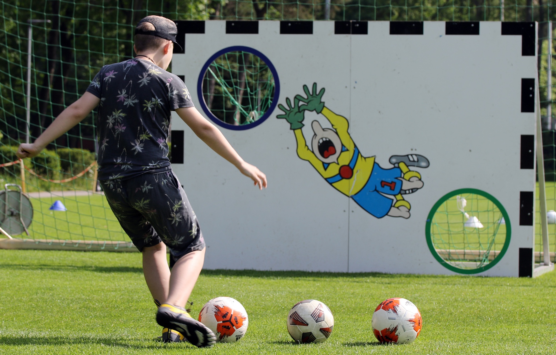 [{"clang_id":1,"value":"Junge schießt Fußball auf ein Zielbrett mit Comic-Torwartabbildung. Drei Bälle stehen bereit. Hintergrund: Fußballtornetz und grüne Bäume."}]