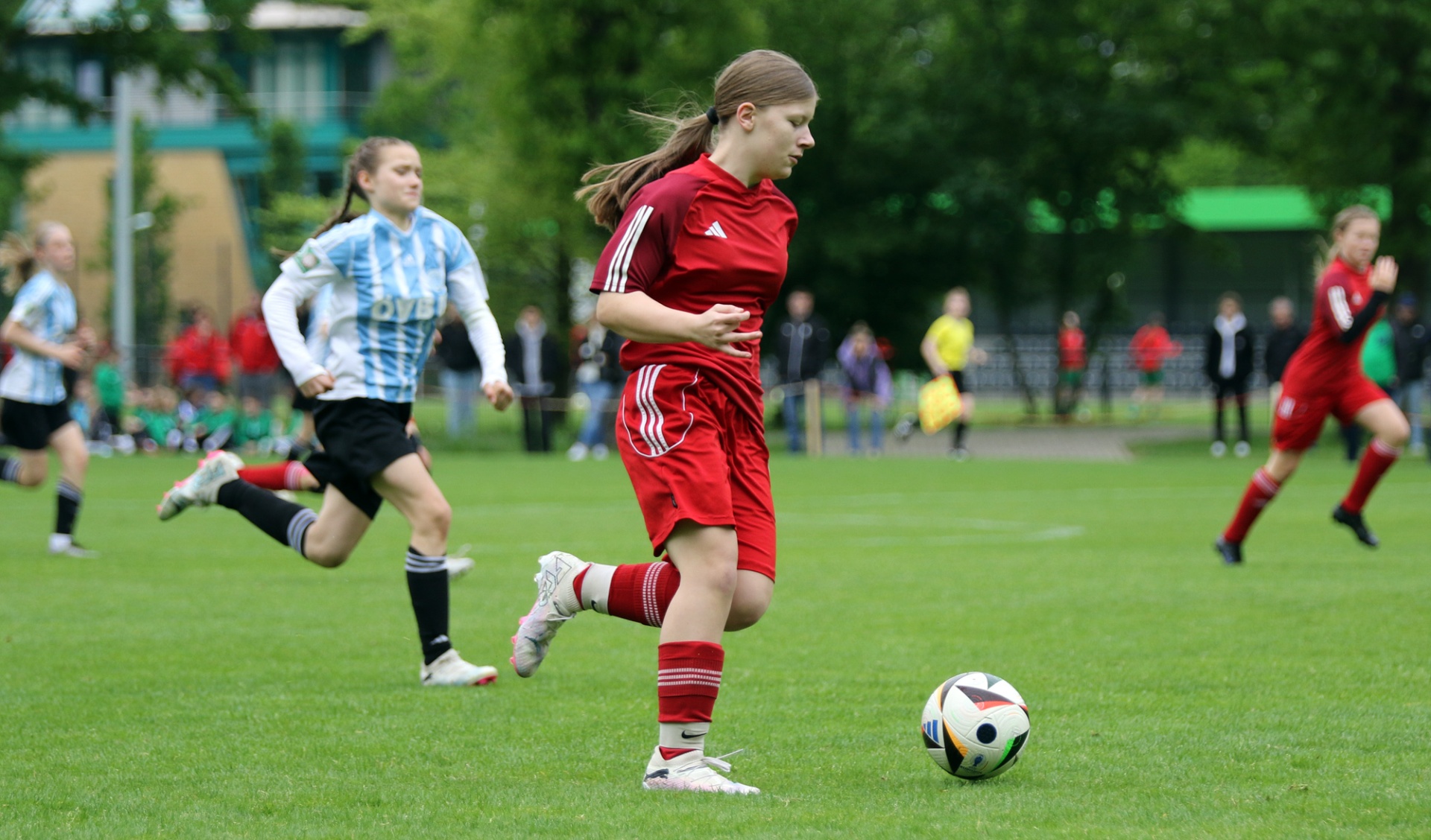 [{"clang_id":1,"value":"Fußballspiel: Junge Spielerin in roten Trikot und Shorts dribbelt den Ball auf grünem Rasen, verfolgt von Gegenspielerinnen. Hintergrund mit Zuschauern und Bäumen."}]