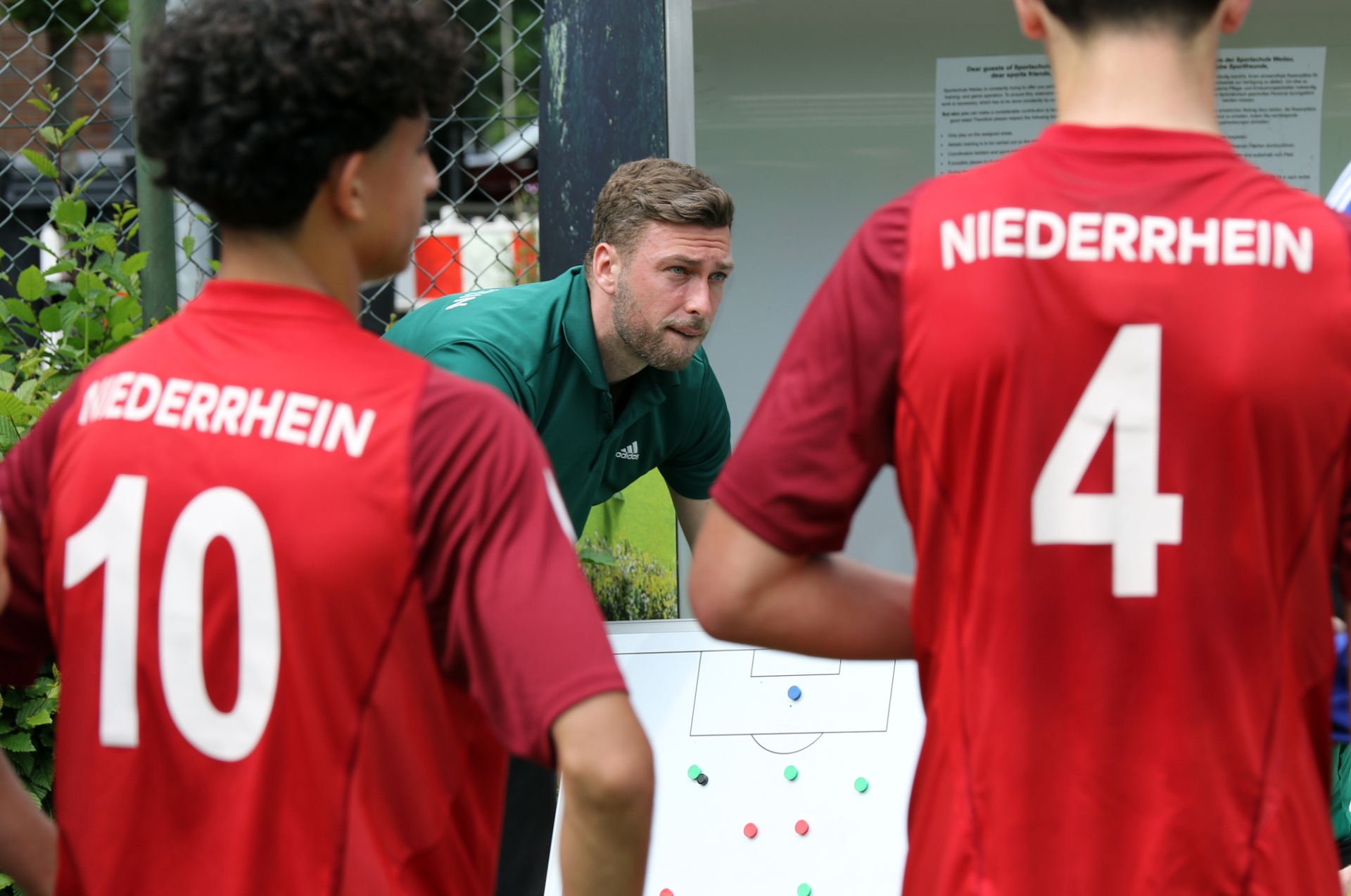 [{"clang_id":1,"value":"Drei Personen beim Fußballtraining. Zwei Spieler mit roten Trikots (Nummern 10 und 4) schauen auf ein taktisches Brett, das ein Trainer erklärt."}]