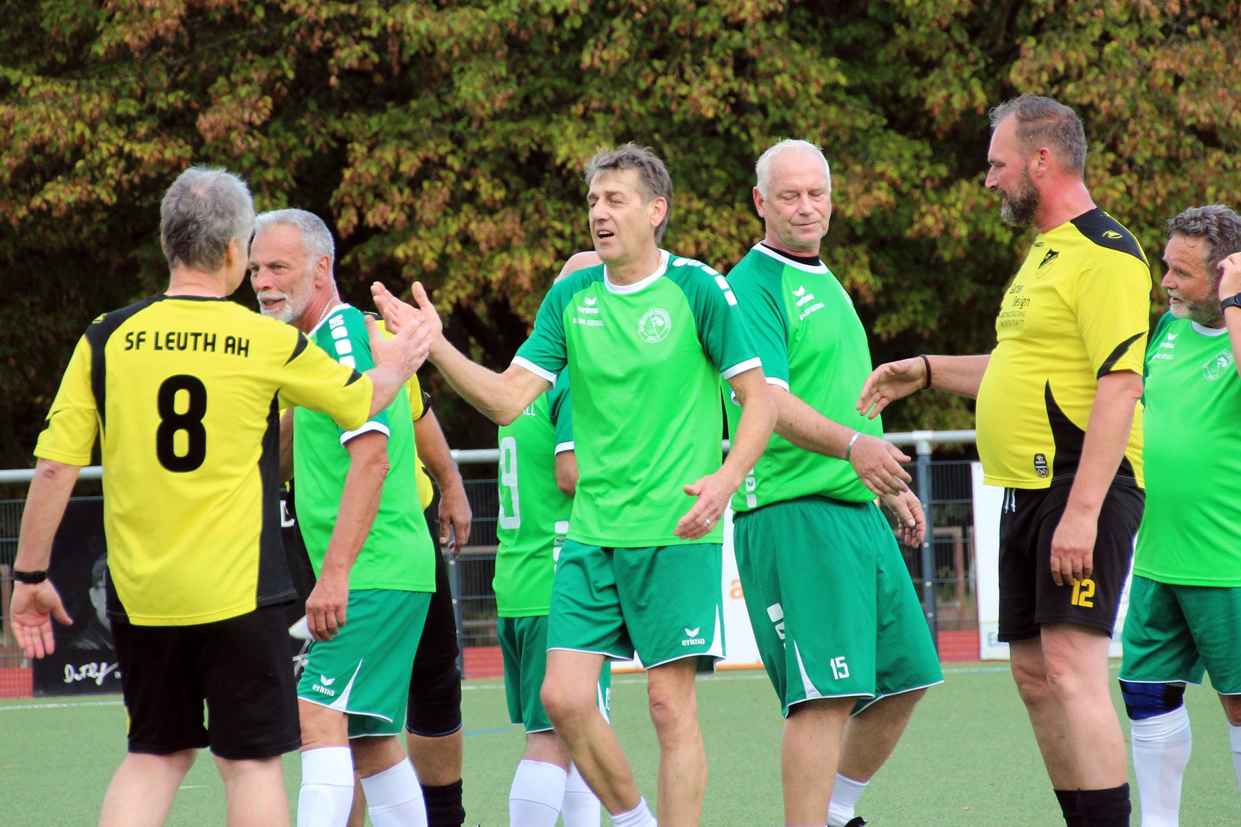 [{"clang_id":1,"value":"Fußballspieler in grünen und gelben Trikots stehen auf einem Rasenplatz und diskutieren. Einige geben sich die Hände. Im Hintergrund Bäume."}]