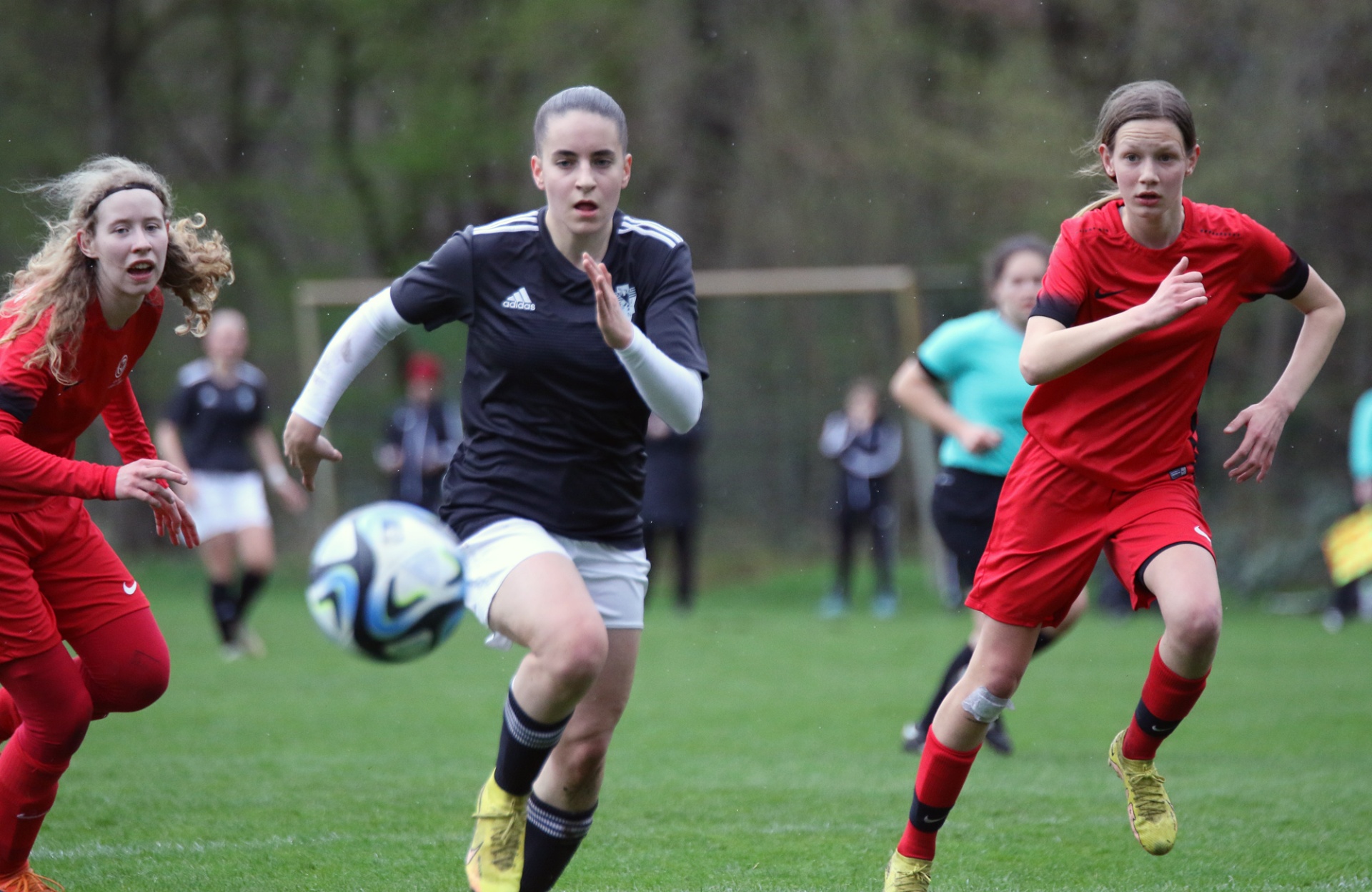[{"clang_id":1,"value":"Drei Fußballspielerinnen auf Grünfeld. Mittlere Spielerin in Schwarz dribbelt Ball, linke in Rot rennt, rechte in Rot folgt. Hintergrund weitere Spieler."}]