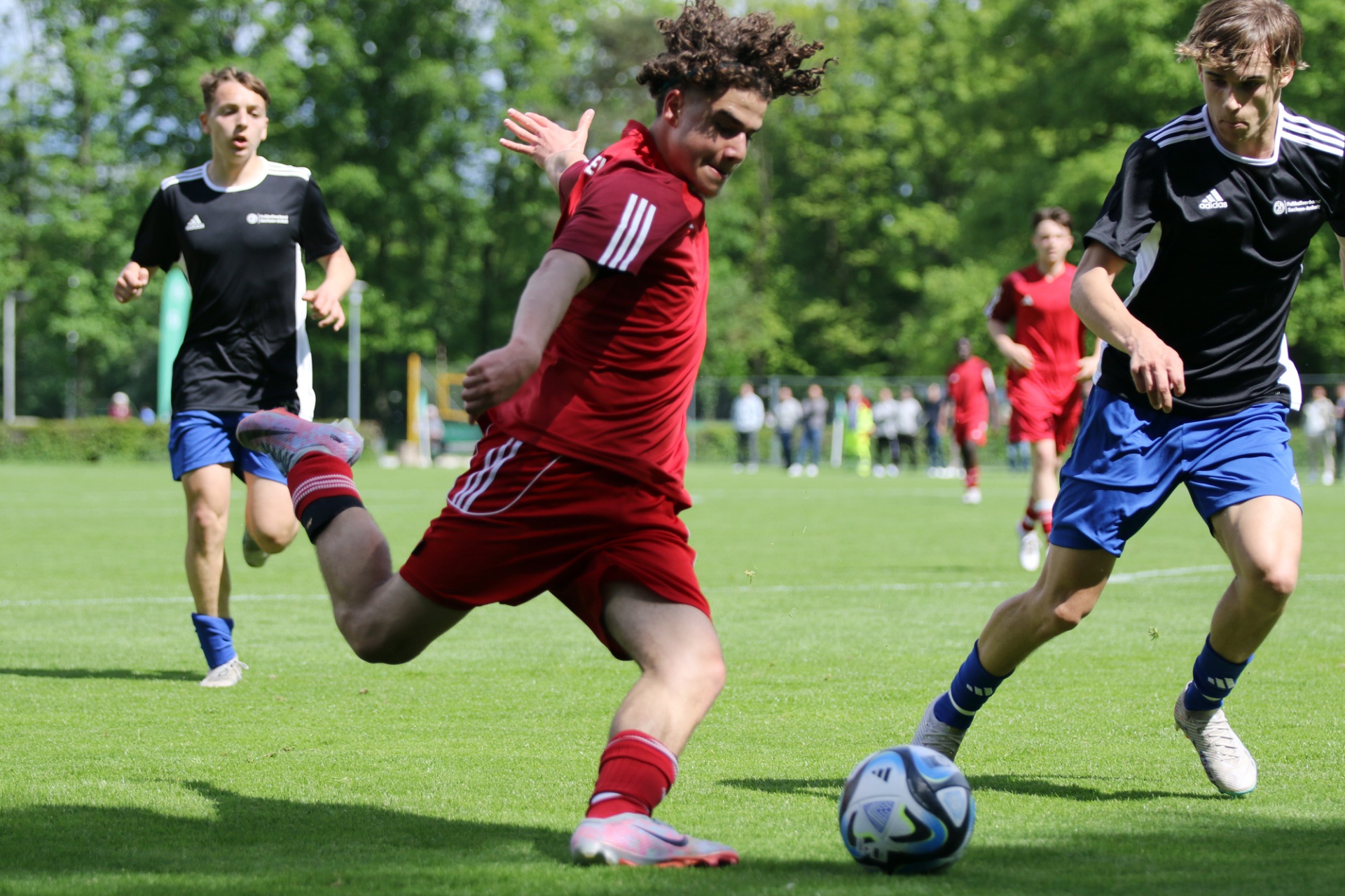 [{"clang_id":1,"value":"Fußballspiel: Spieler in Rot schießt auf das Tor, zwei weitere Spieler in Schwarz-Weiß und Blau stehen in der Nähe. Grüner Rasenplatz, Hintergrund mit Bäumen."}]