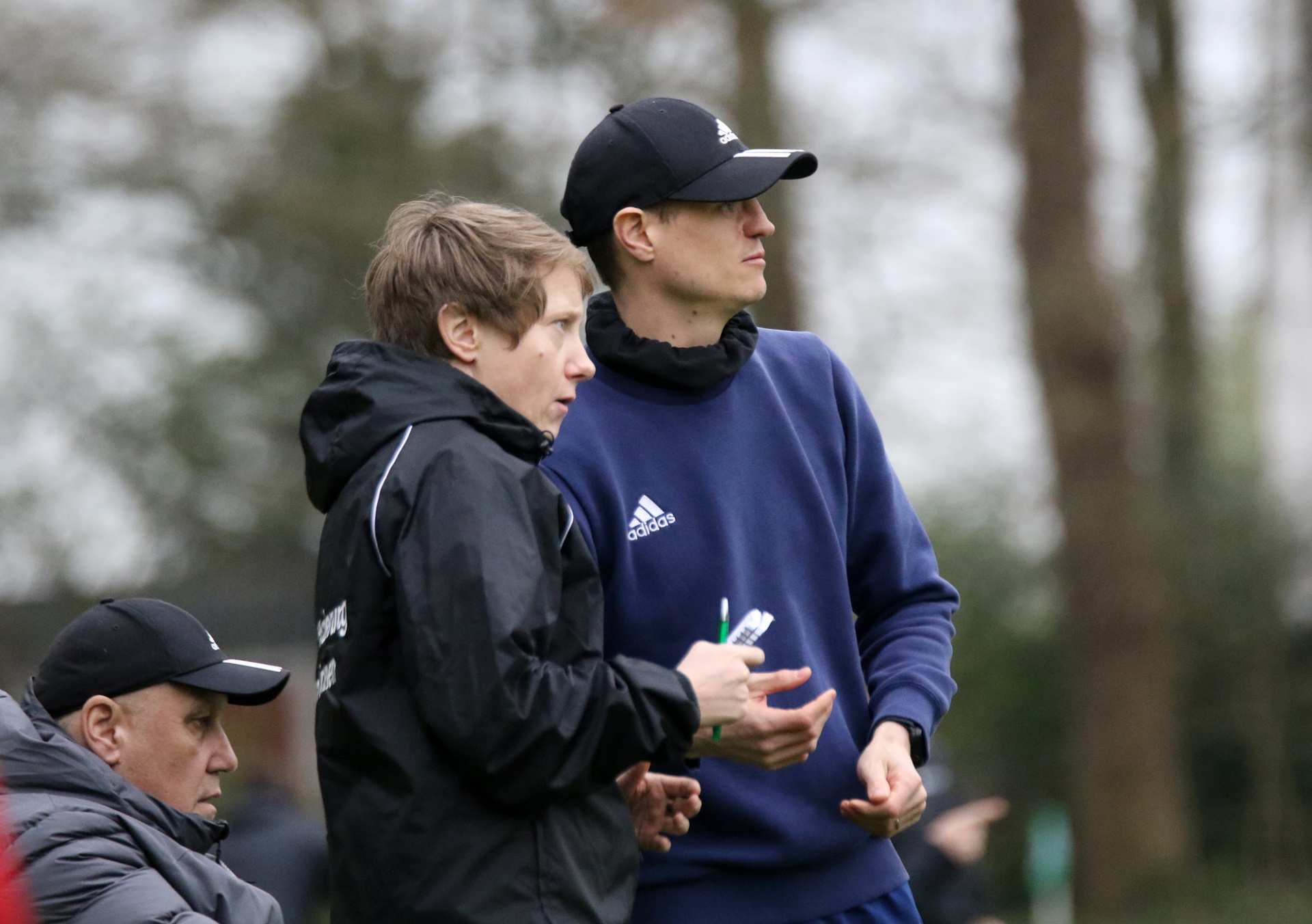 [{"clang_id":1,"value":"Drei Männer mit Baseballkappen und Sportbekleidung stehen auf einer grünen Wiese vor Bäumen. Zwei diskutieren angeregt, der dritte blickt zur Seite."}]