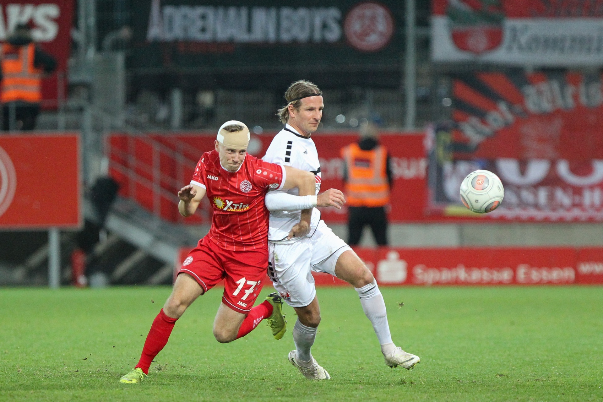 [{"clang_id":1,"value":"Zwei Fußballspieler im Wettkampf: Links in Rot, rechts in Schwarz-Weiß. Im Hintergrund Banner und Zuschauerränge. Rasenplatz bei Nacht."}]