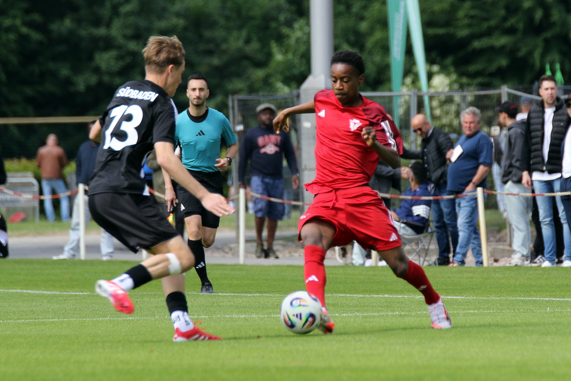 [{"clang_id":1,"value":"Fußballspiel: Spieler in Rot dribbelt vor Gegenspieler in Schwarz. Im Hintergrund Zuschauer und Bäume."}]
