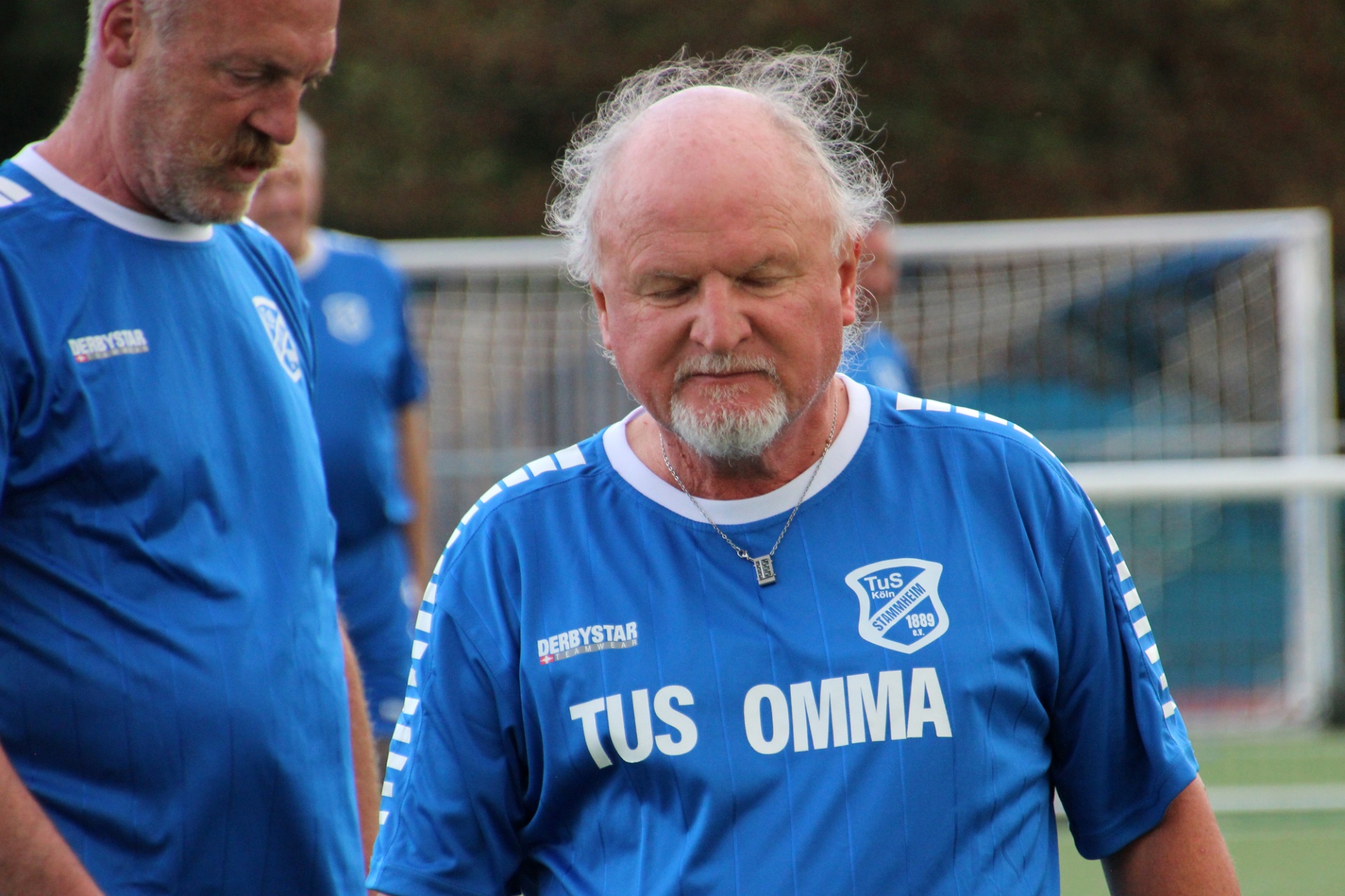 [{"clang_id":1,"value":"Zwei Männer in blauen Fußballtrikots stehen auf einem Sportplatz. Der Mann im Vordergrund hat eine Brille, Bart und ist kahlköpfig. Im Hintergrund ein Fußballtor."}]