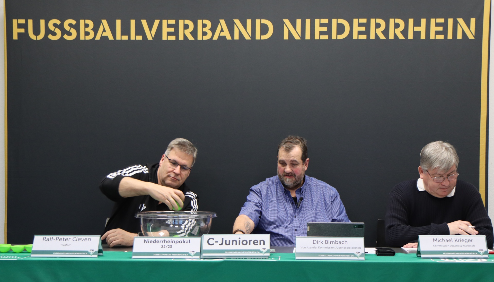 [{"clang_id":1,"value":"Drei Männer sitzen an einem Tisch mit Namensschildern vor einem Banner mit dem Schriftzug \"FUSSBALLVERBAND NIEDERRHEIN C-Junioren\". Ein Pokal steht auf dem Tisch."}]