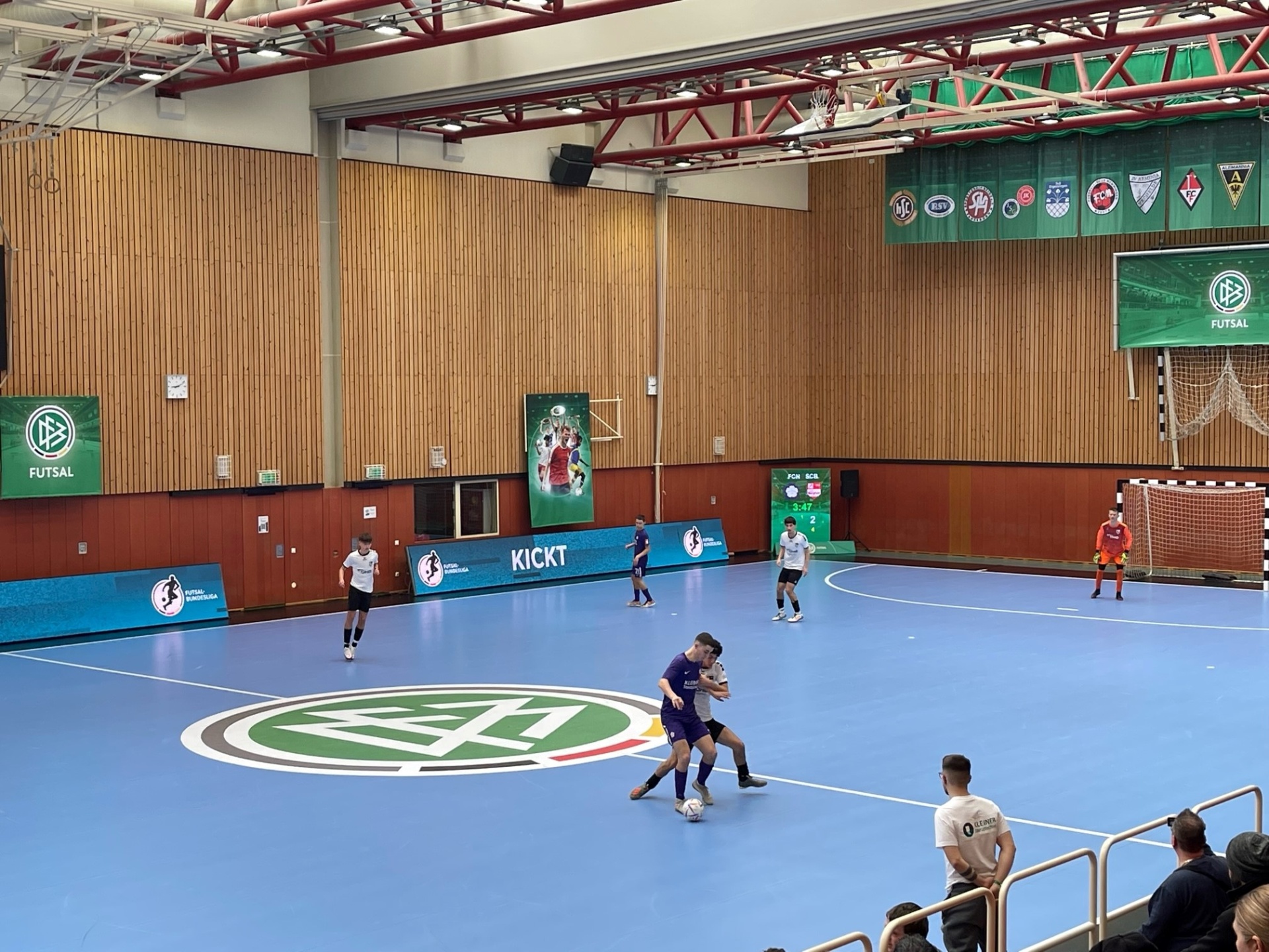 [{"clang_id":1,"value":"Futsal-Halle mit laufendem Spiel. Spieler in blauen und weißen Trikots auf blauem Feld mit Kreismarkierung. Zuschauerreihen im Hintergrund."}]