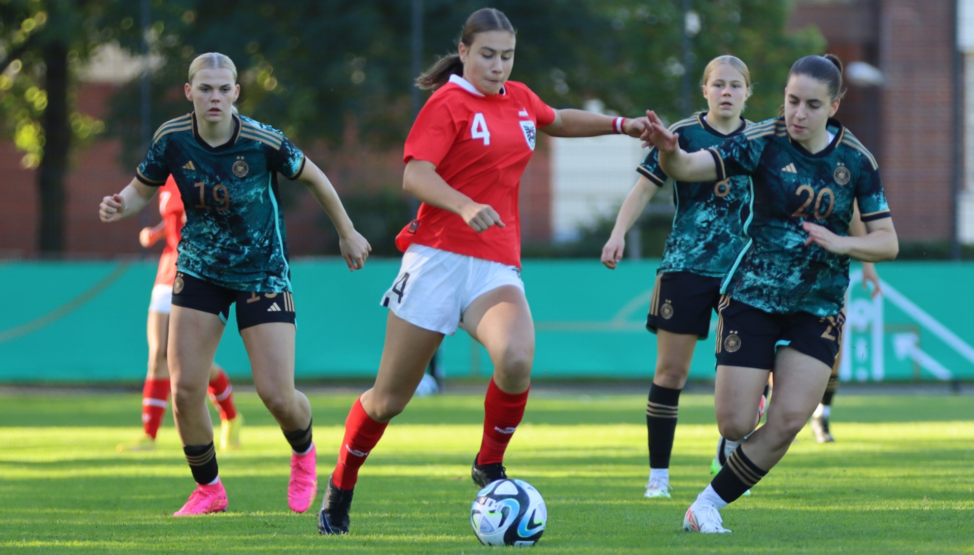[{"clang_id":1,"value":"Fußballspiel: Spielerin mit rotem Trikot und Nummer 4 dribbelt den Ball, verfolgt von drei Spielerinnen in dunkelgrünen Trikots mit Nummern 7, 16 und 20. Rasenplatz im Hintergrund."}]