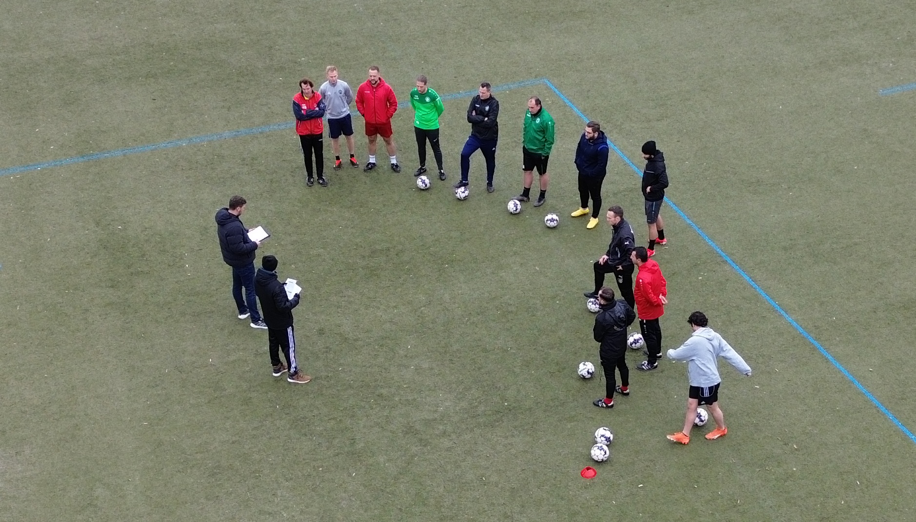[{"clang_id":1,"value":"Fußballteam im Kreis auf grünem Kunstrasenfeld. Trainer mit Schreibblock erklärt Taktik. Bälle verteilt im Kreis."}]