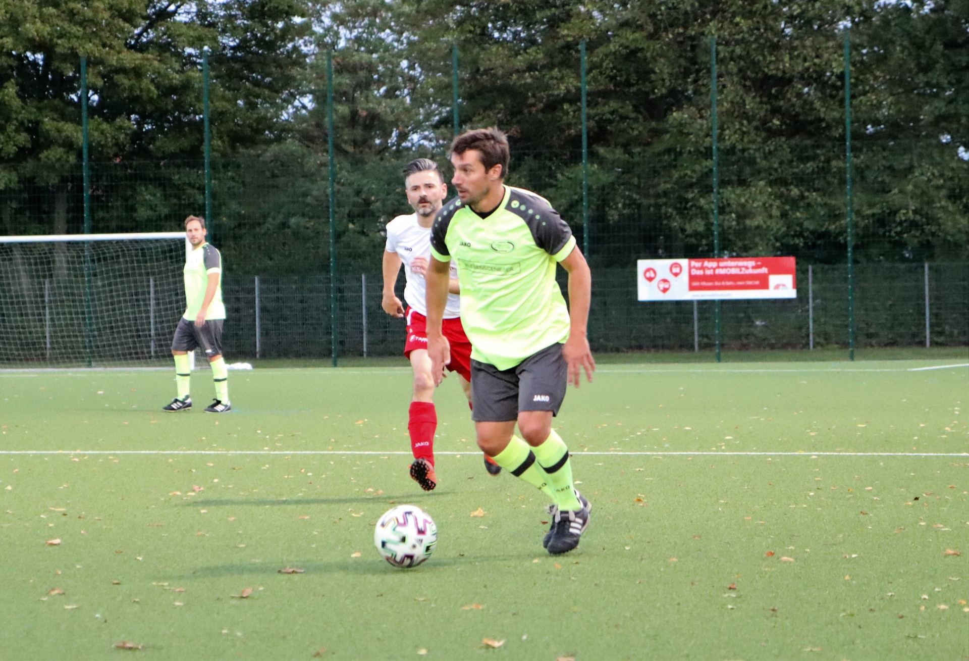[{"clang_id":1,"value":"Fußballspiel: Spieler mit grünem Trikot dribbelt Ball auf grünem Kunstrasen. Im Hintergrund weitere Spieler und ein Zaun."}]