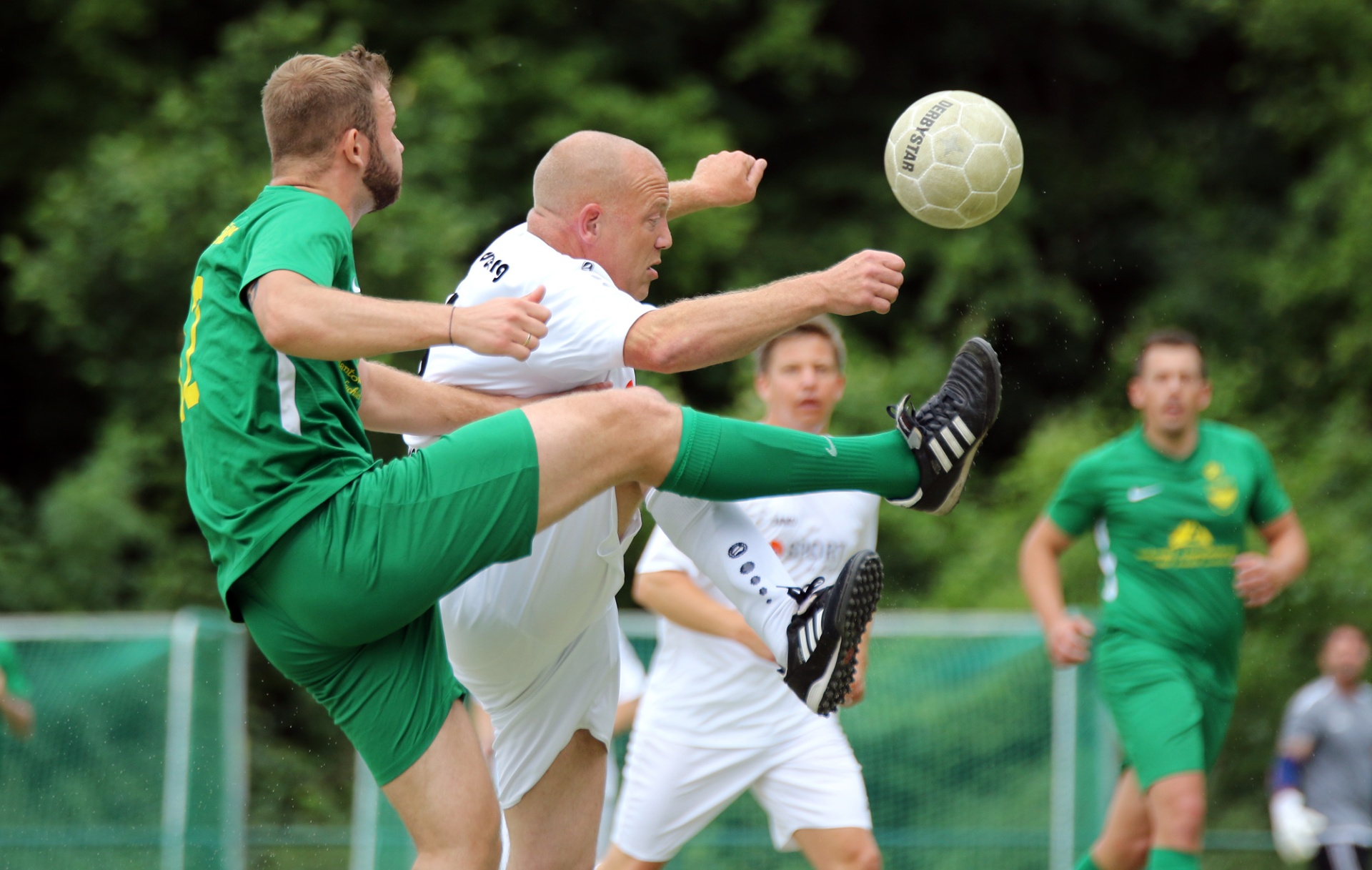 [{"clang_id":1,"value":"Zwei Fußballspieler kämpfen um den Ball. Links trägt einer grünes Trikot, rechts weißes. Im Hintergrund weitere Spieler und ein Zaun."}]