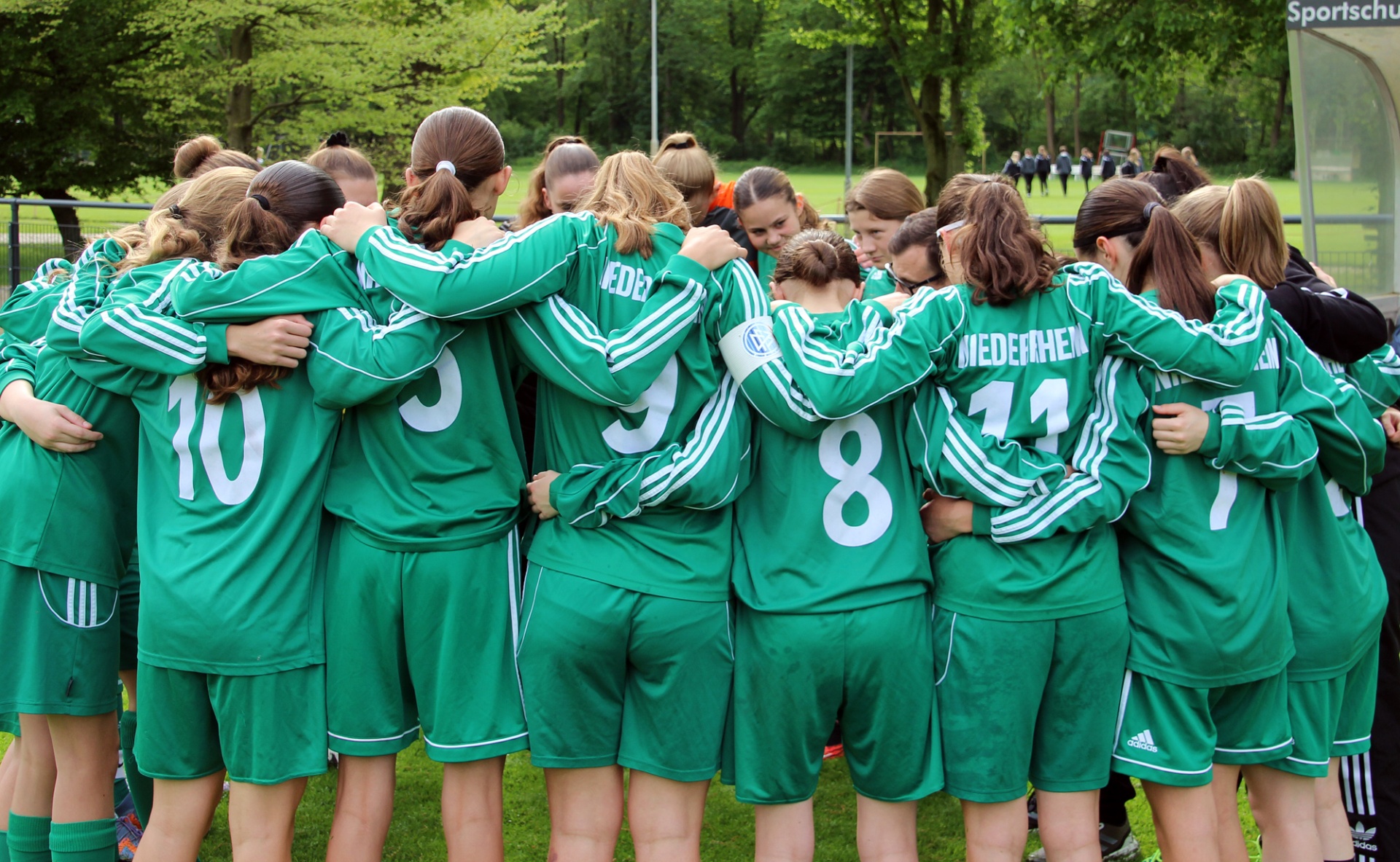 [{"clang_id":1,"value":"Fußballmannschaft in grünen Trikots und Shorts im Kreis stehend, Arme um die Schultern. Nummern auf den Rückseiten sichtbar. Hintergrund: Sportplatz und Bäume."}]