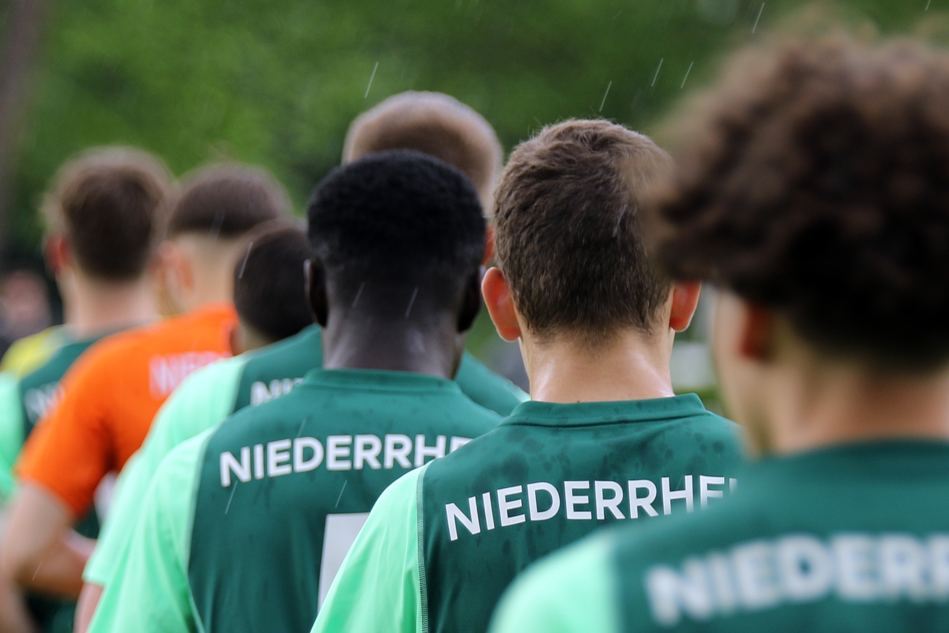 [{"clang_id":1,"value":"Fußballmannschaft in grünen Trikots mit Aufschrift \"NIEDERRHEIN\" steht auf einem Fußballfeld. Rückansicht der Spieler."}]