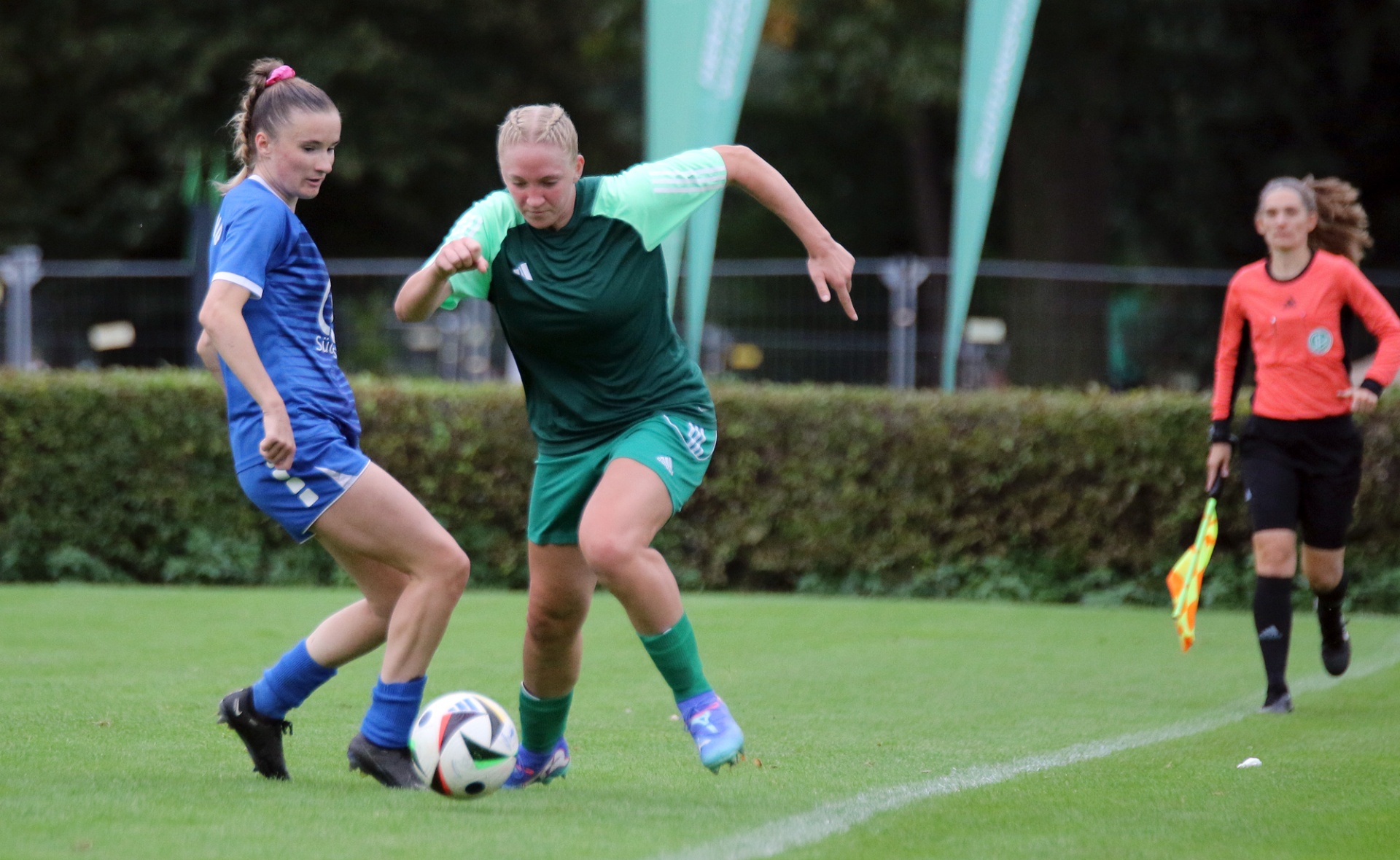[{"clang_id":1,"value":"Zwei Fußballspielerinnen kämpfen um den Ball auf grünem Rasen. Links in blauem Trikot, rechts in grünem Trikot mit hellgrünen Ärmeln. Im Hintergrund eine Schiedsrichterin."}]