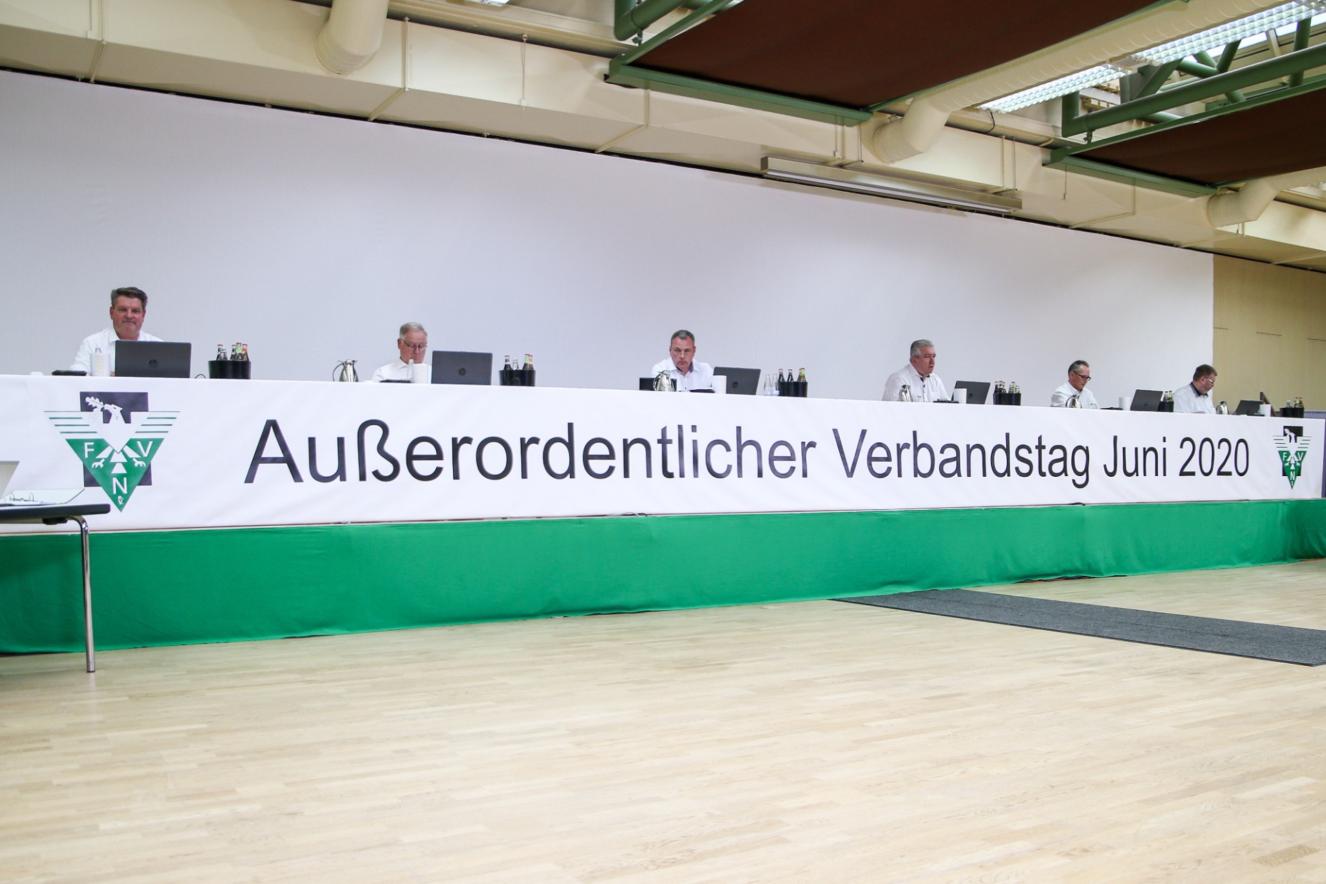 [{"clang_id":1,"value":"Reihe von Männern am Konferenztisch mit grünem Banner \"Außerordentlicher Verbandstag Juni 2020\". Großer heller Raum mit hölzernem Boden."}]